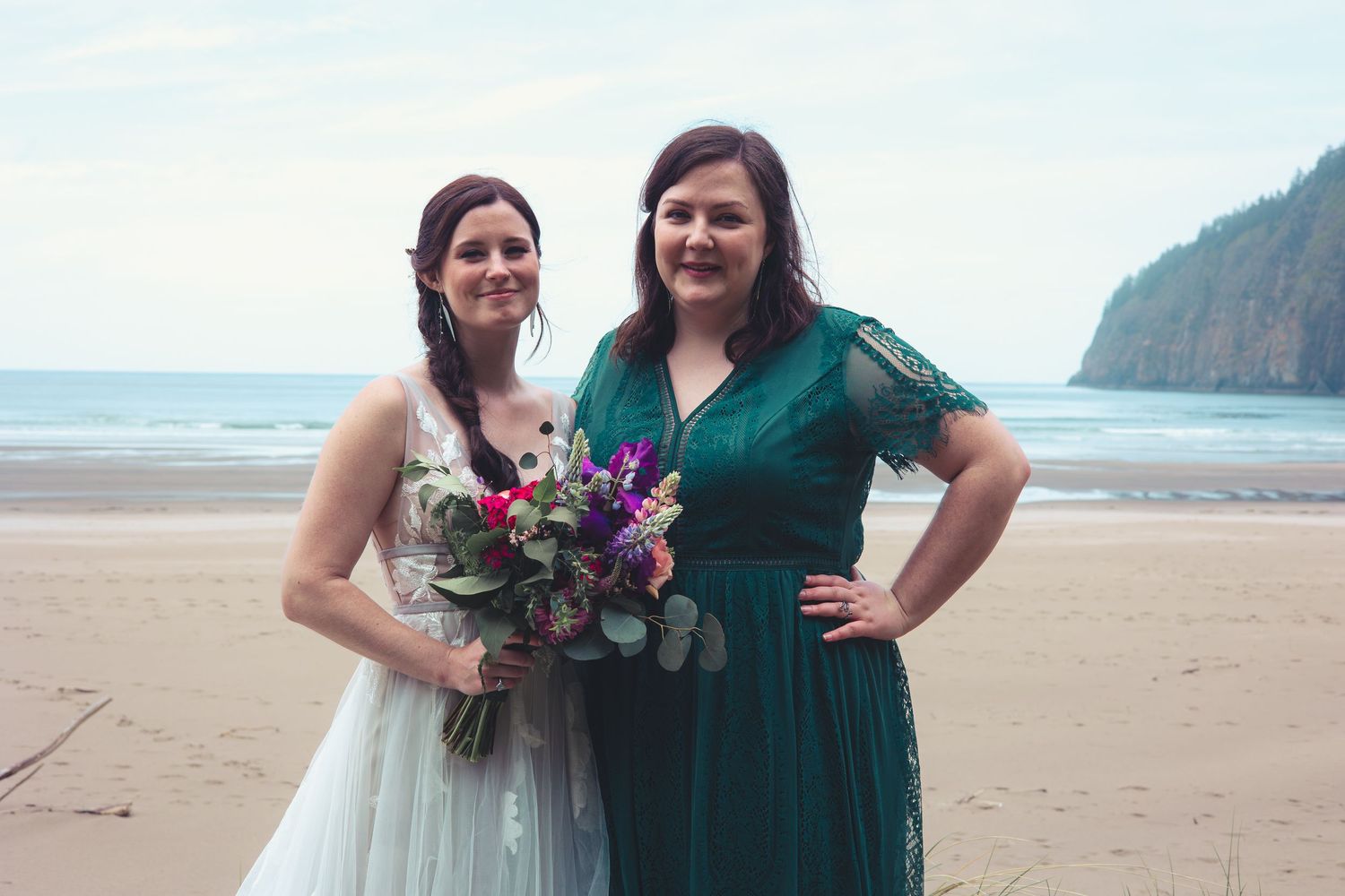 A bride holding a colorful bouquet and her bridesmaid in a teal dress smile on a scenic beach.