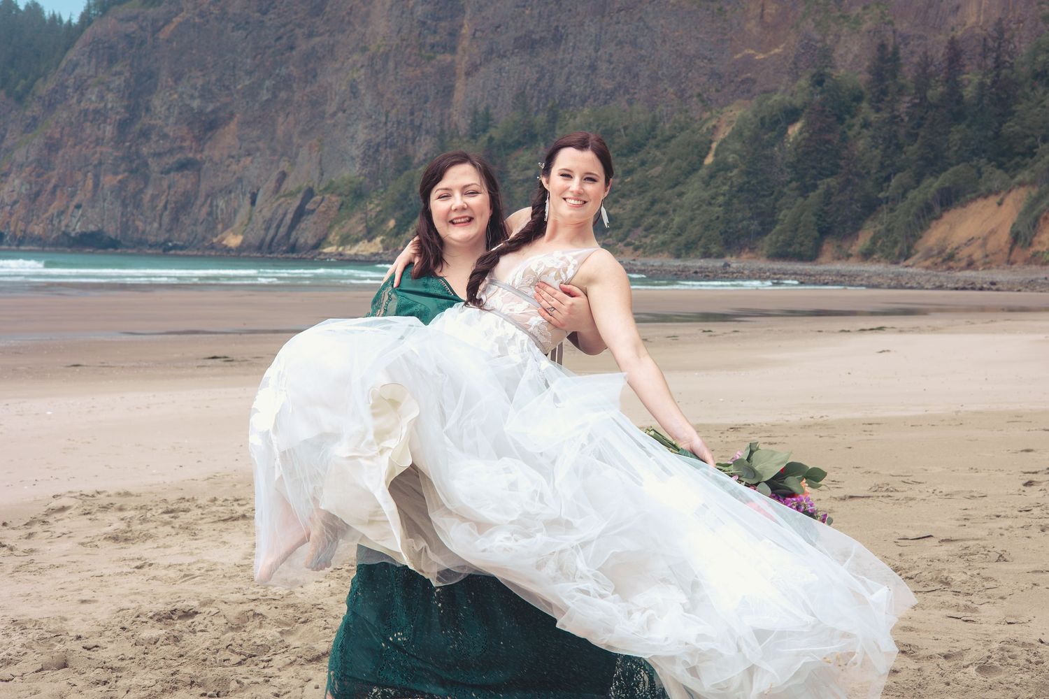 Woman in green dress carries laughing bride in white gown on a scenic sandy beach.
