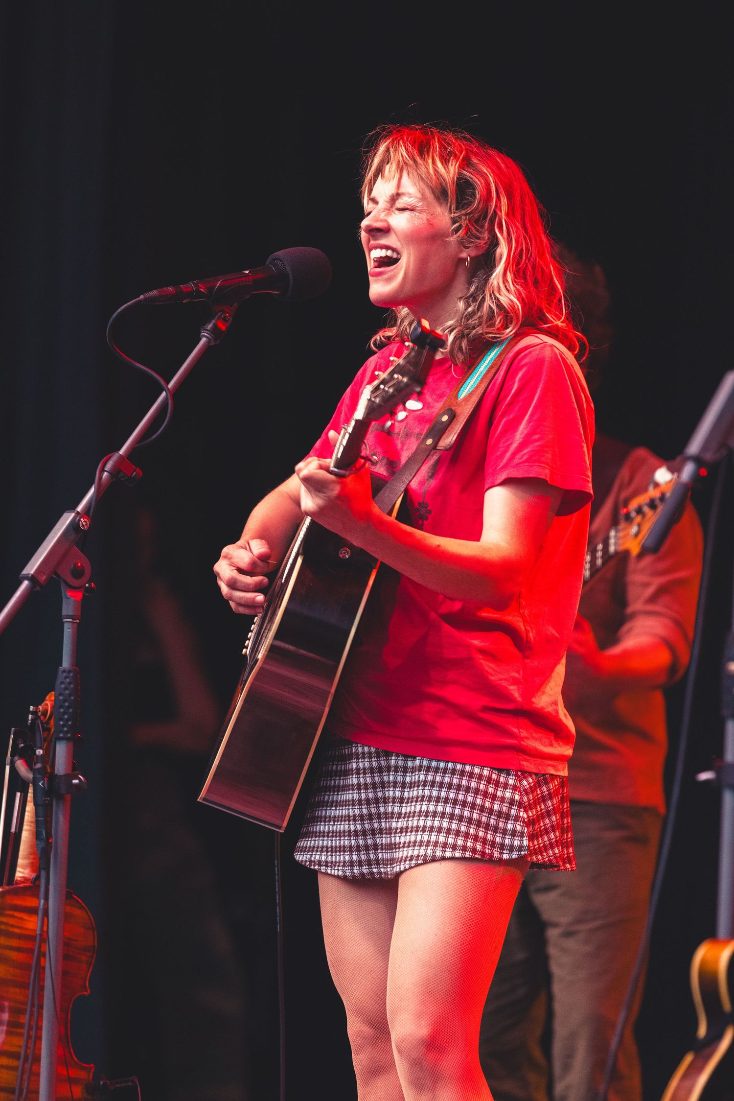Woman in red shirt joyfully singing and playing acoustic guitar under red stage lights.