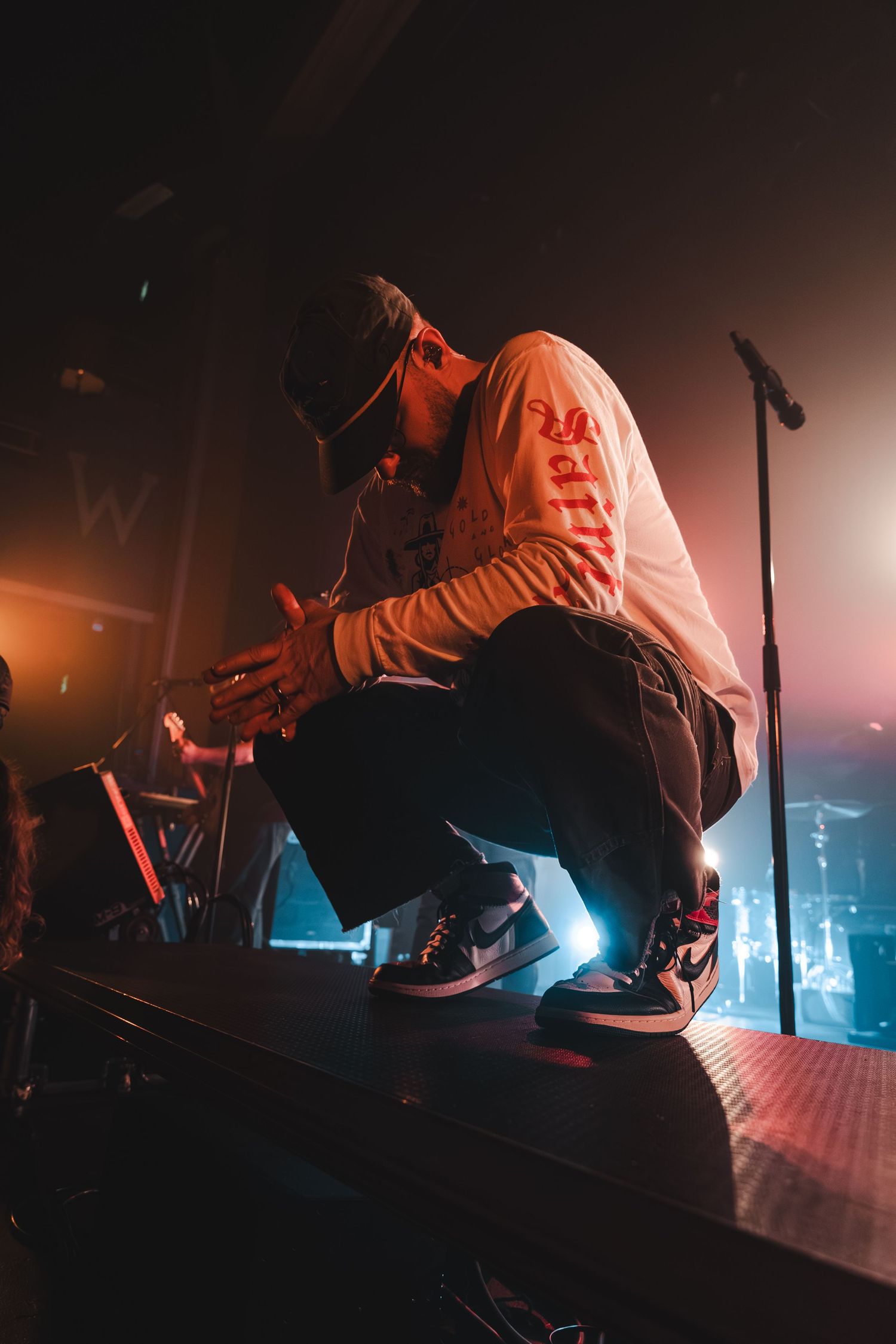 Male performer crouching on stage edge in Nike sneakers during live concert.