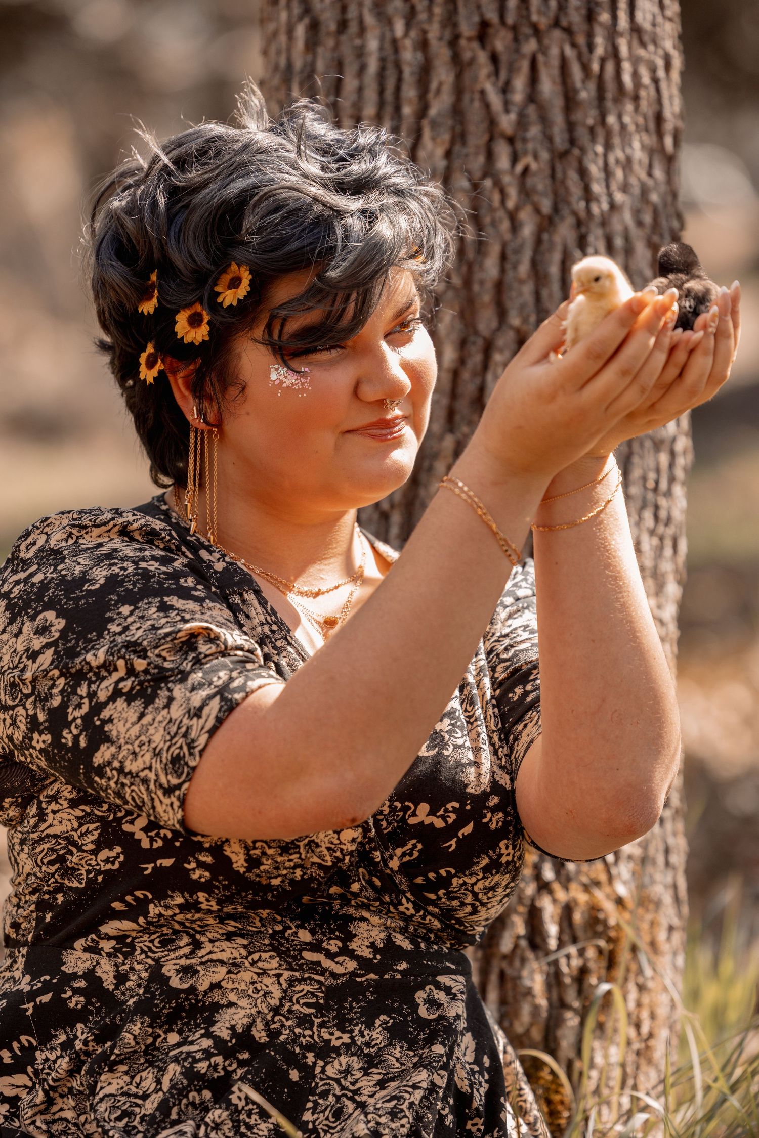 Young woman with floral hair clips gently cradles a baby chick in cupped hands beside a tree.