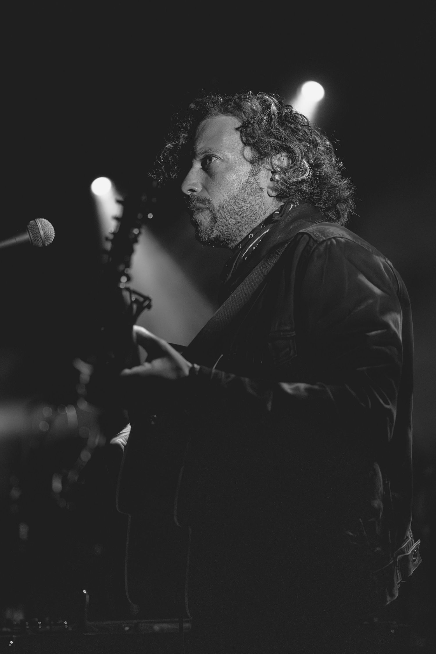 Black and white portrait of curly-haired bearded guitarist performing on stage near microphone at live concert.