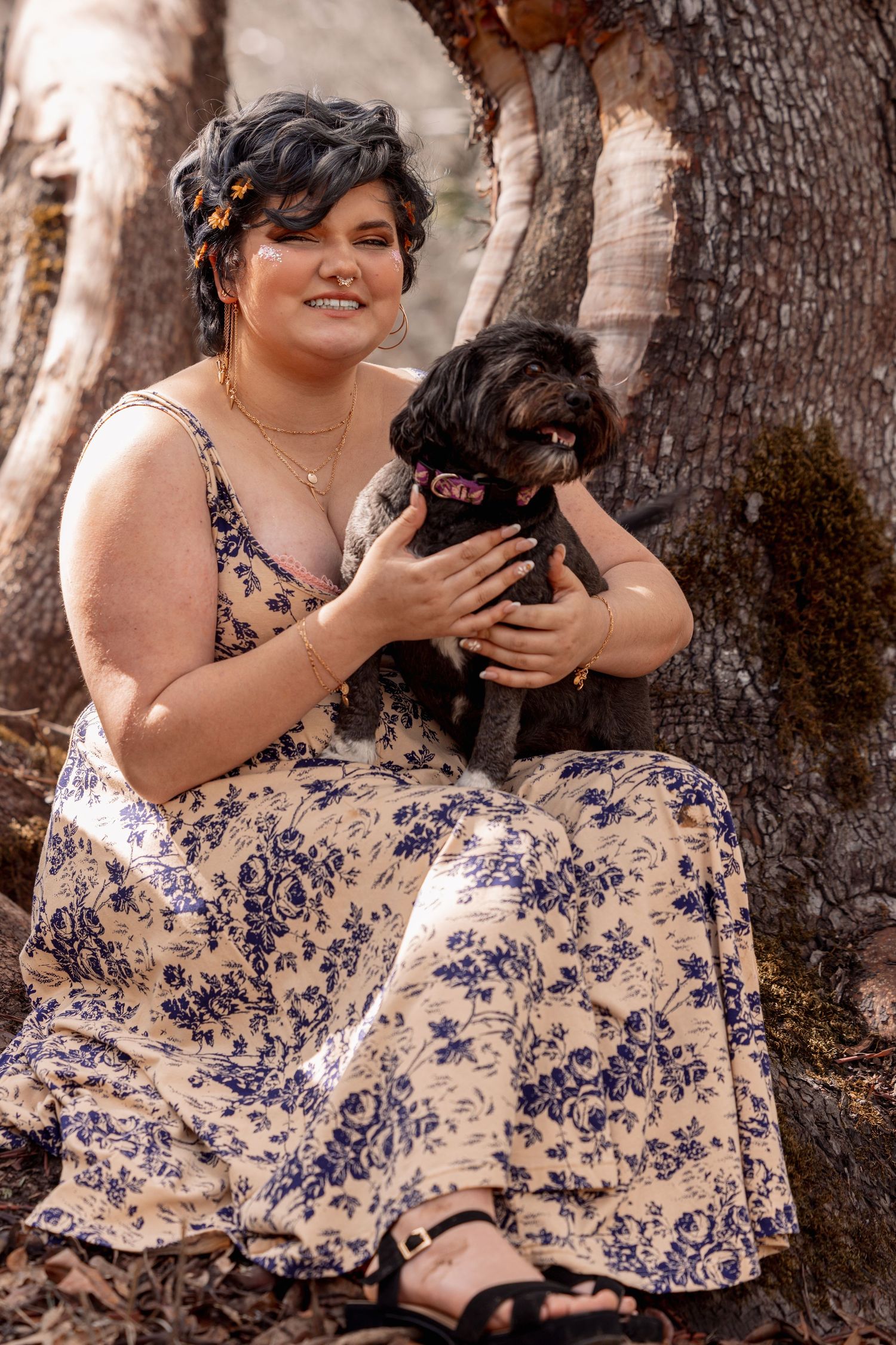 Smiling woman in floral dress sitting against tree trunk holding small black dog.