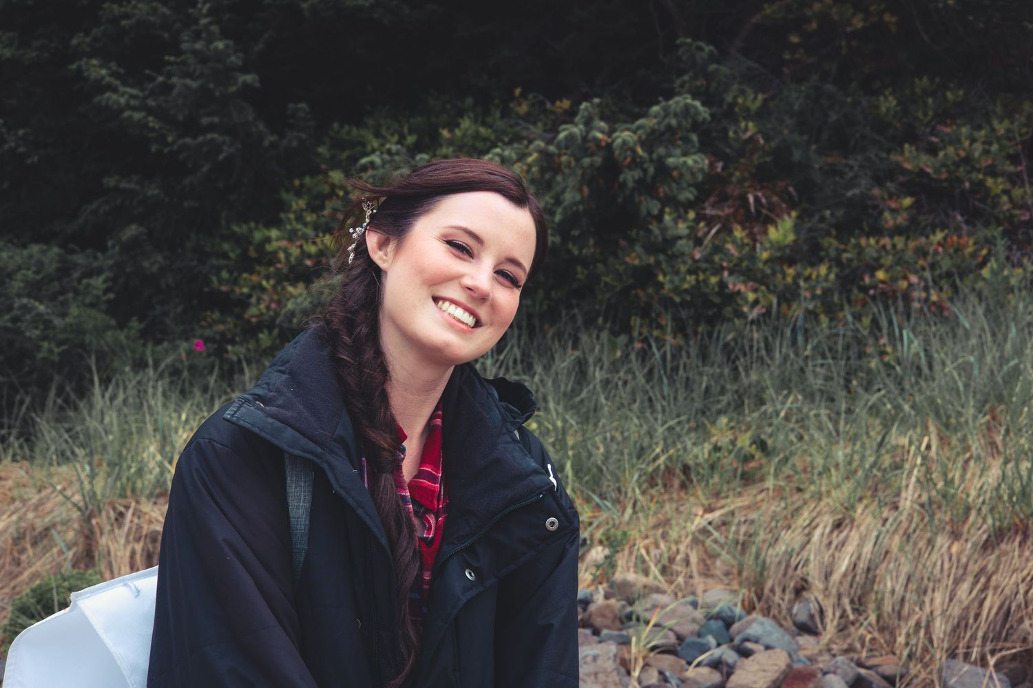 Smiling woman with braided hair in a dark jacket sitting on rocks near a grassy outdoor area.