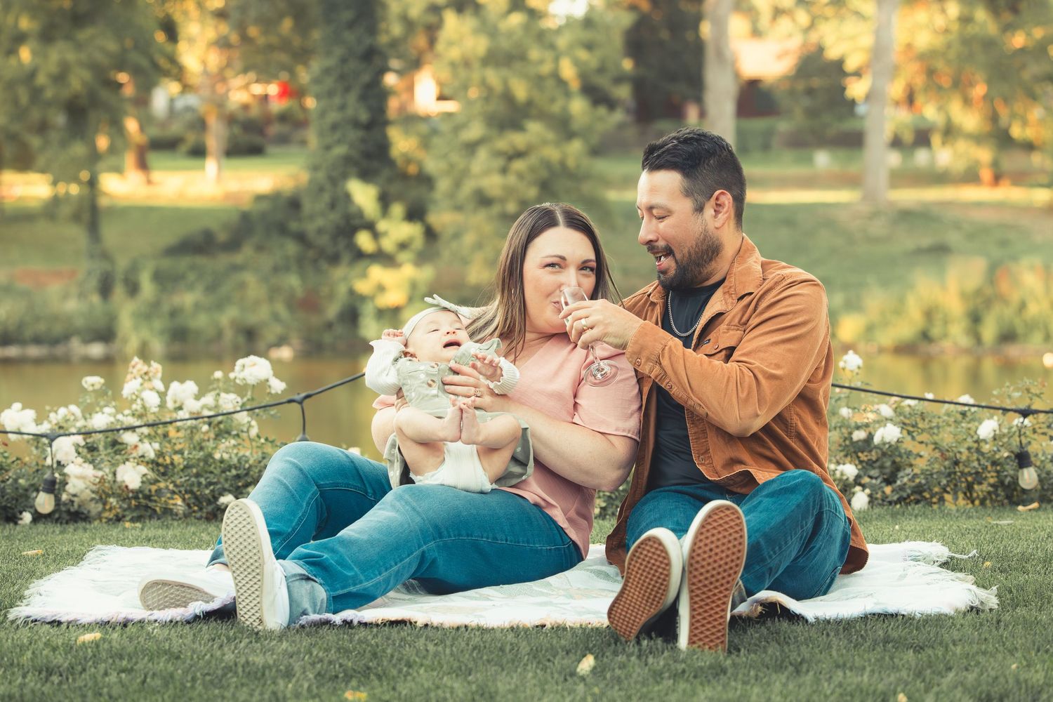 Couple laughing and bonding with their baby on a white blanket outdoors.
