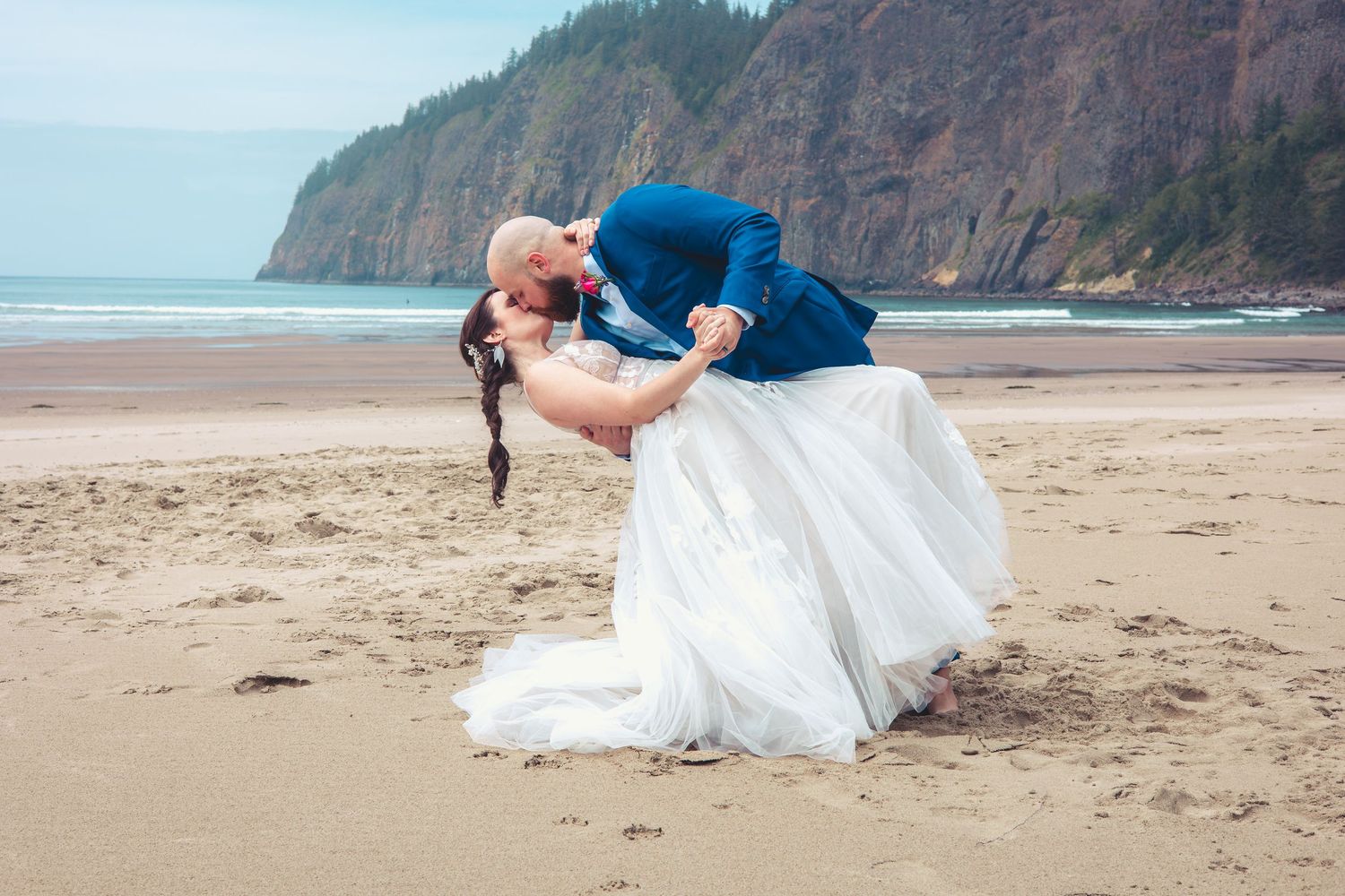 Groom dips bride for a romantic kiss on sandy beach, her gown sweeping the sand, cliffs in background.