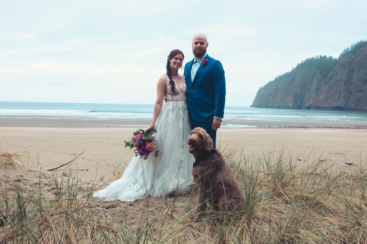 Bride in white gown and groom in blue suit stand with dog on sandy beach.