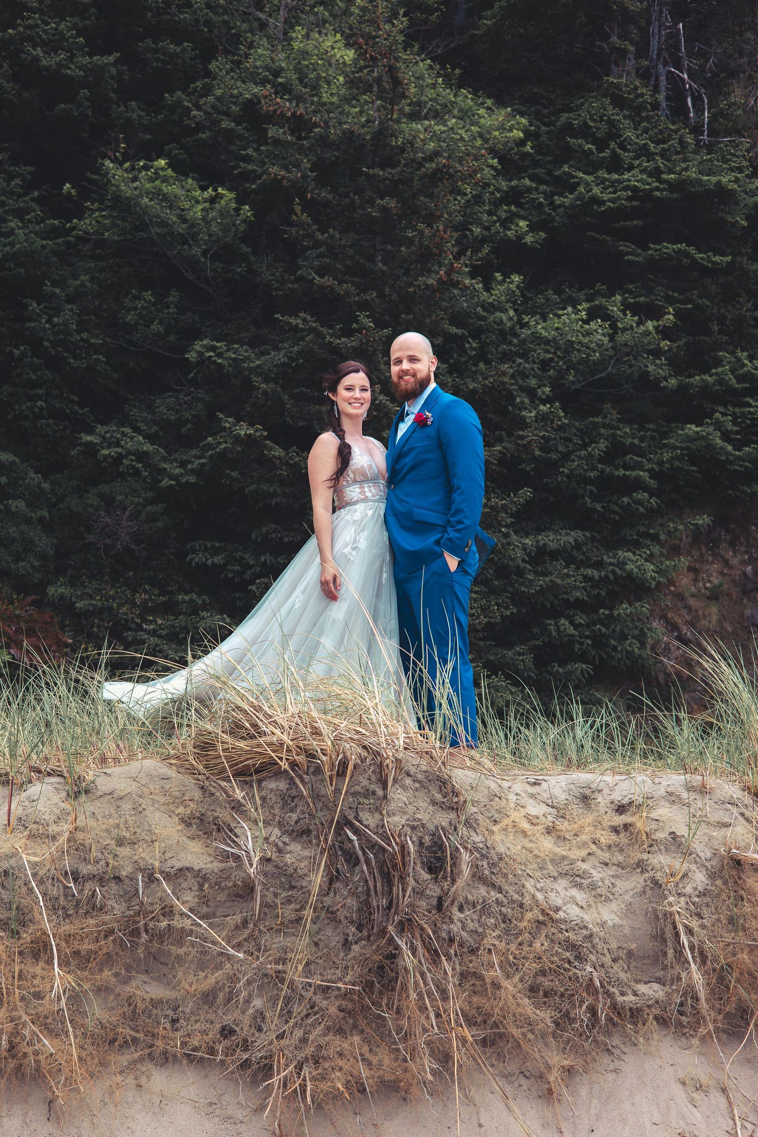 Wedding couple poses on sandy dune with evergreen forest backdrop at beach elopement.