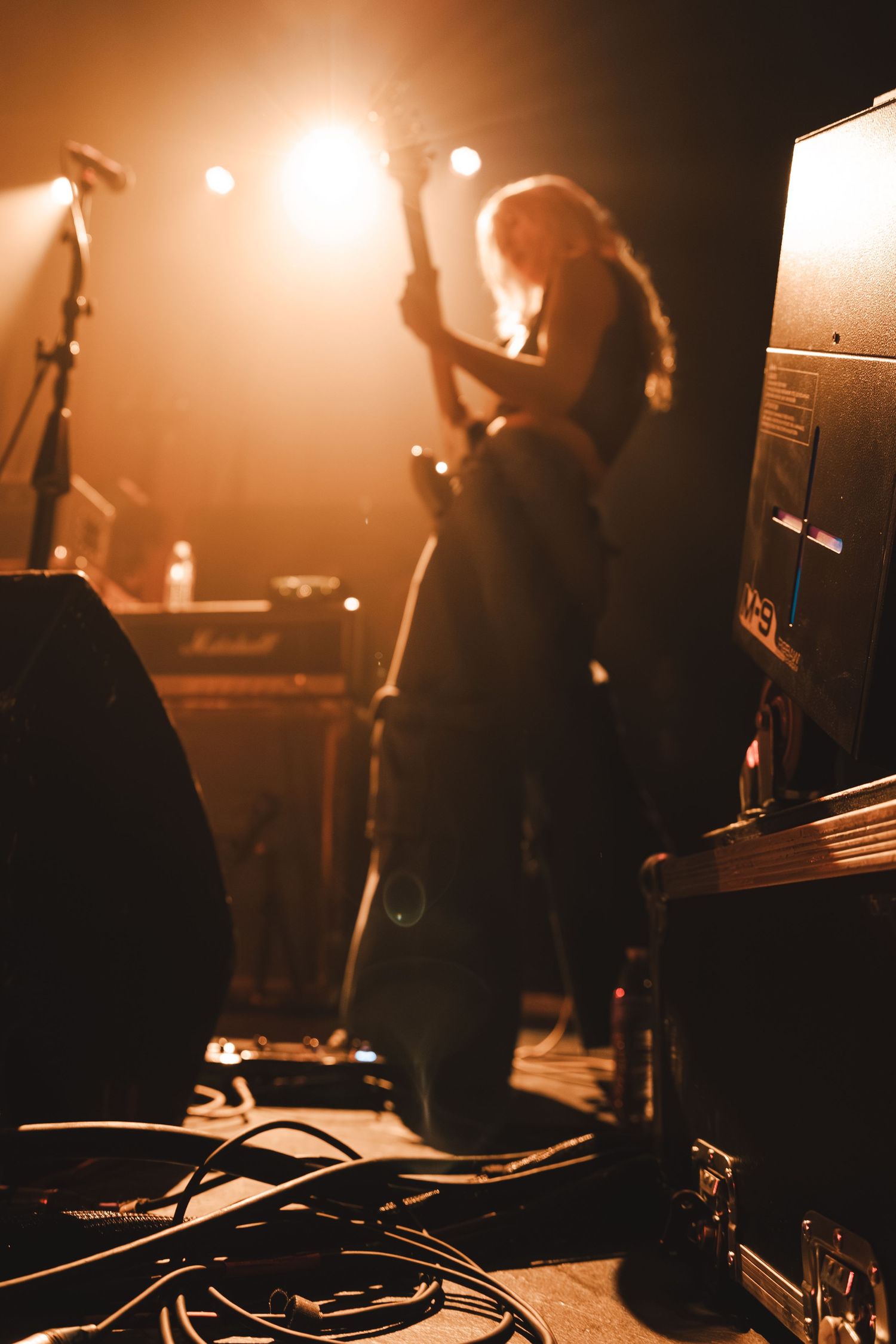 Silhouette of blonde guitarist performing on stage surrounded by Marshall amplifiers and stage cables.