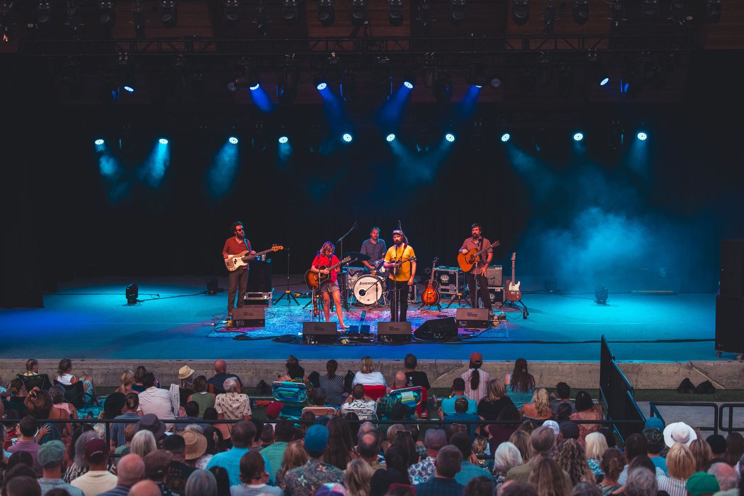 Five-piece band performing on a lit stage with blue and purple lights before a large indoor concert audience.