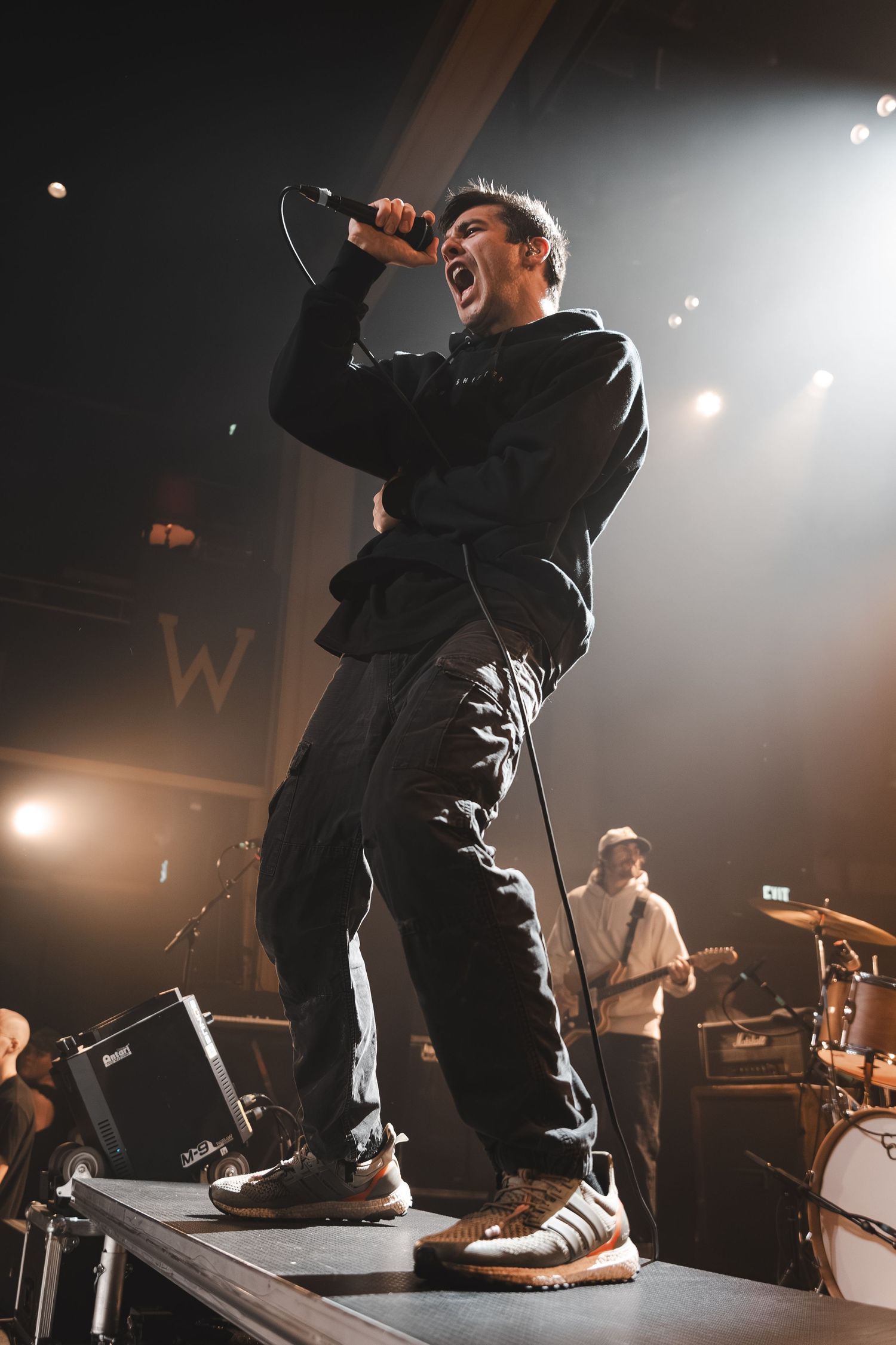 Male vocalist in black hoodie sings passionately into microphone on stage with dramatic stage lighting behind him.