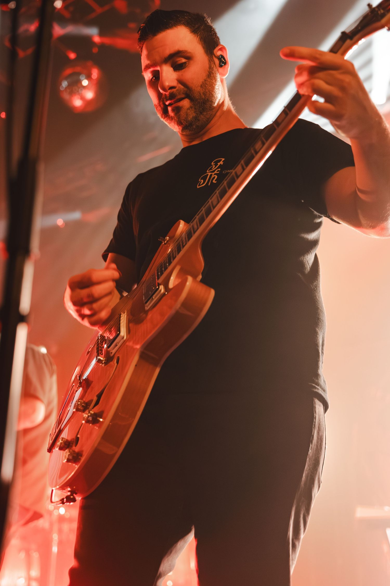 Bearded guitarist smiling while playing electric guitar on stage under red lights.