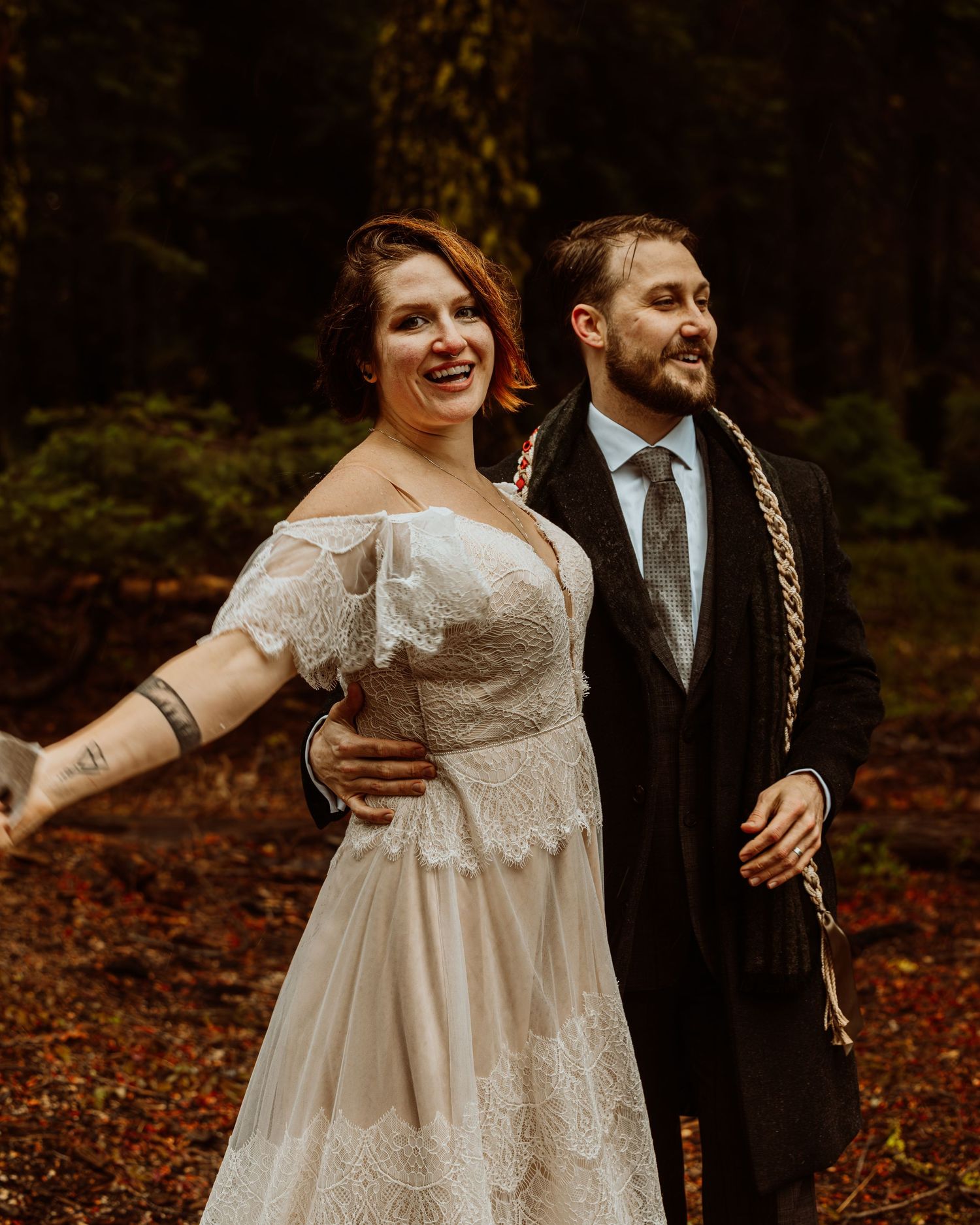 Smiling bride in lace off-shoulder gown and groom in suit walking together in an autumn forest.