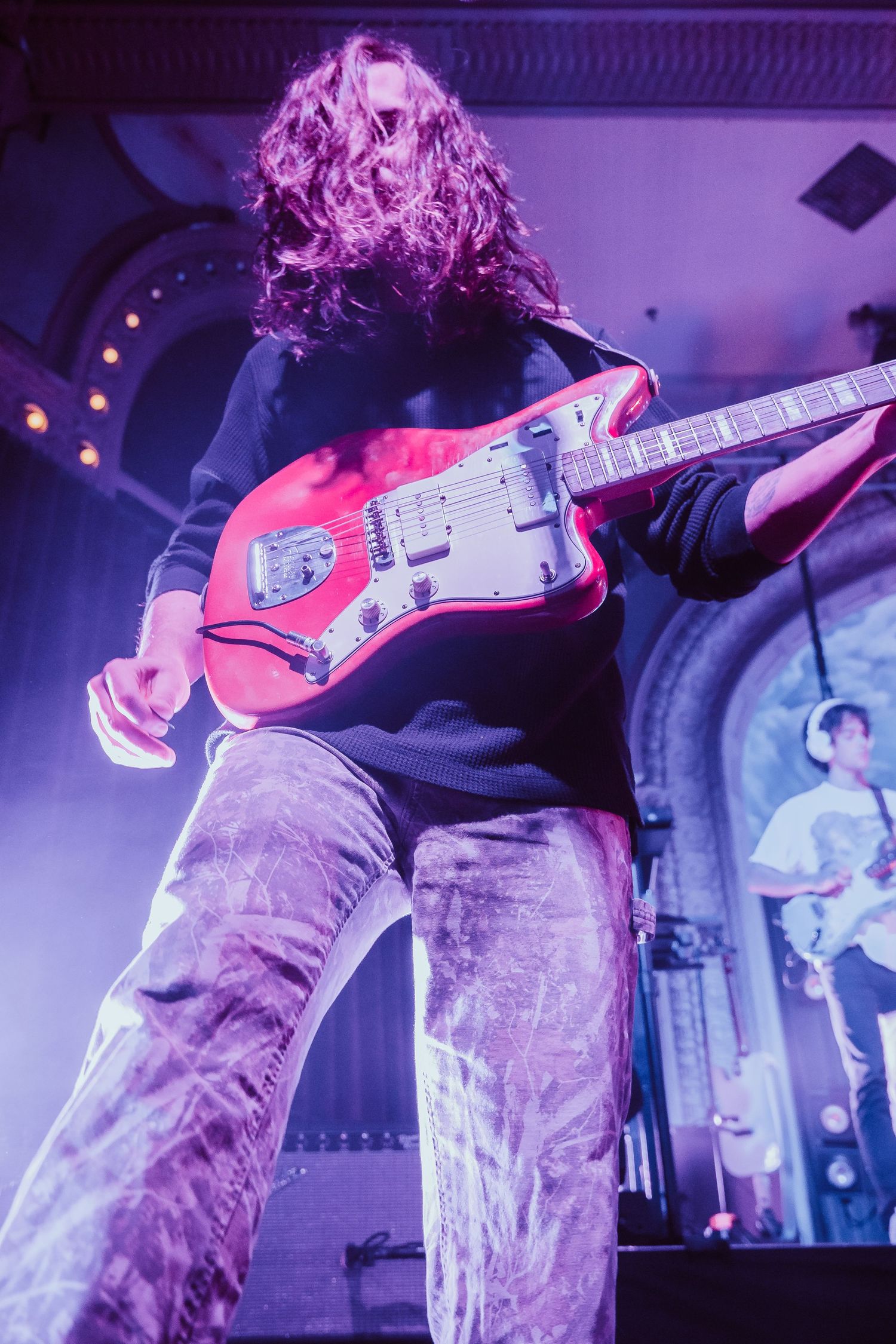 Low-angle view of curly-haired guitarist playing red Jazzmaster guitar on a purple-lit stage.