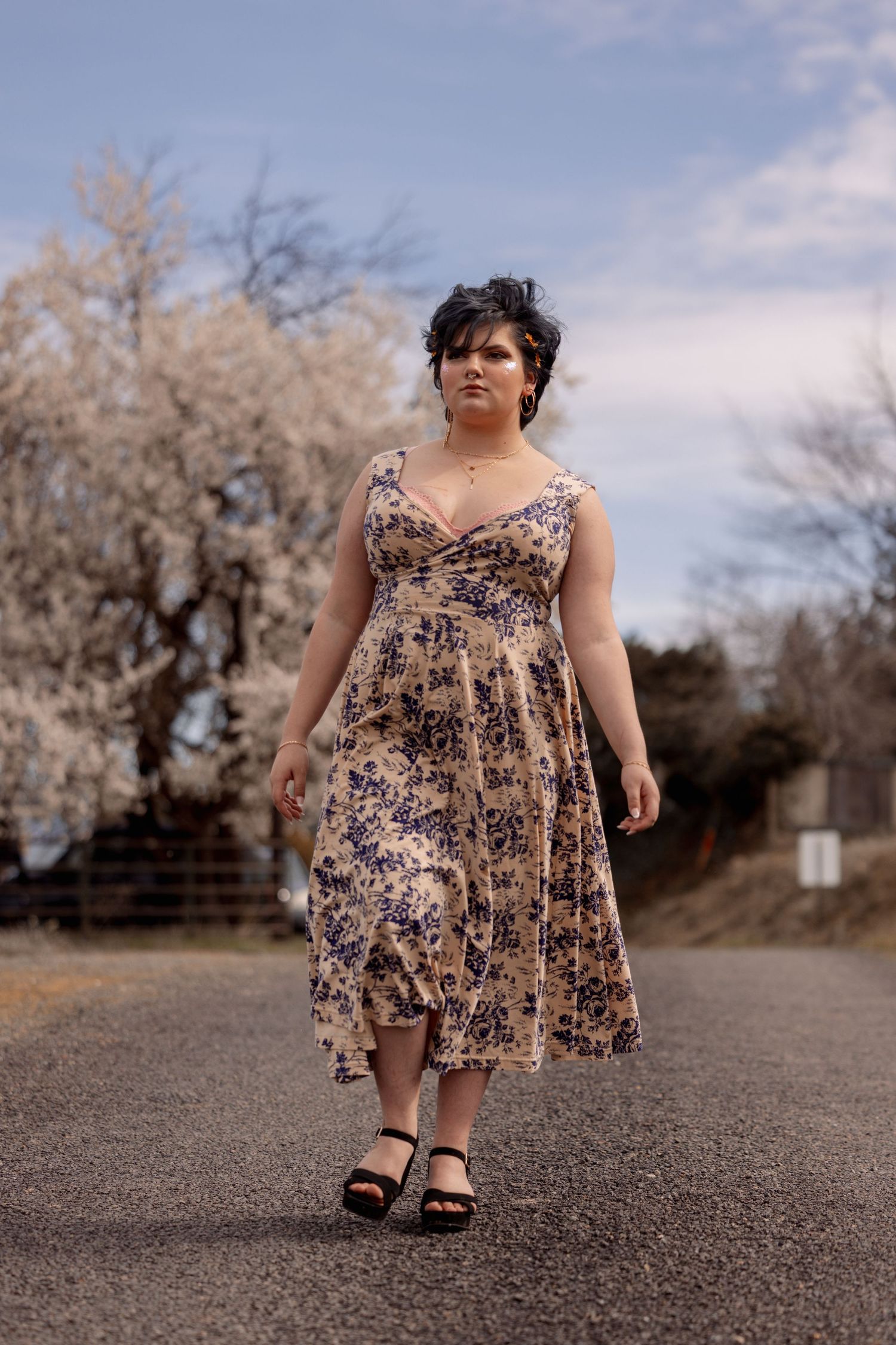 Woman in beige blue floral dress walking on country road flanked by spring flowering trees.
