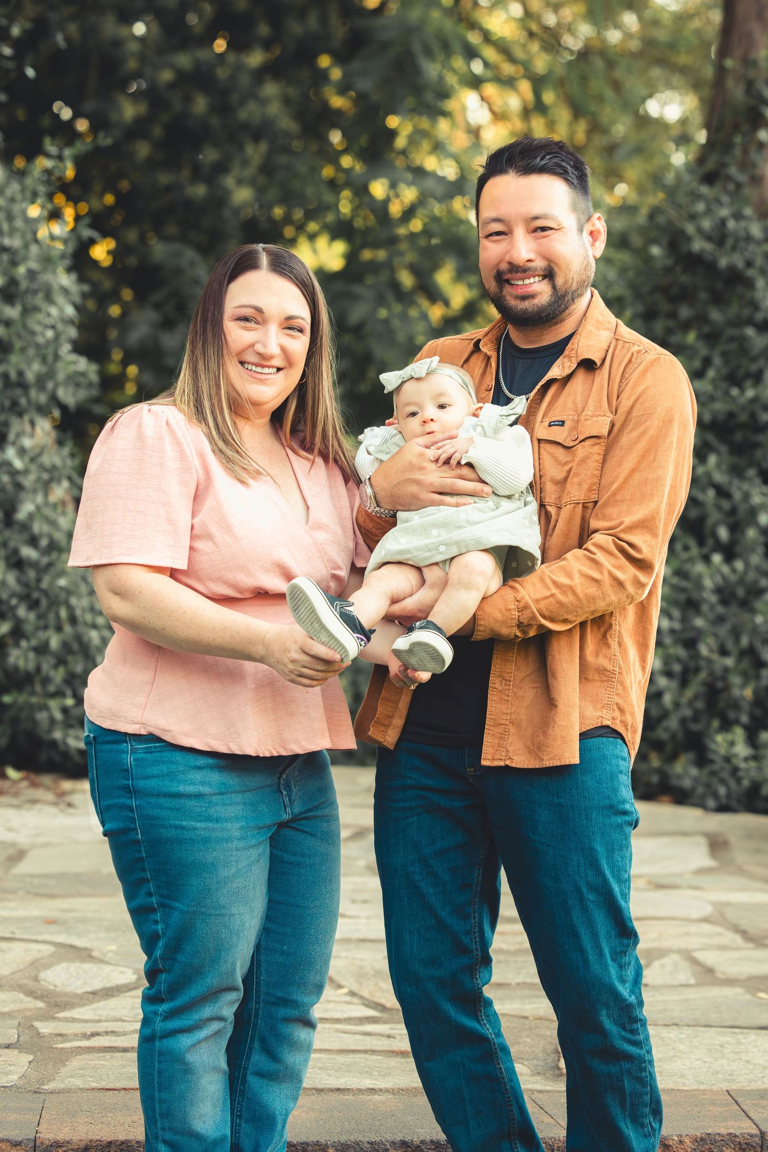 Smiling family of three standing outdoors, father holding baby girl in green dress with green foliage background.