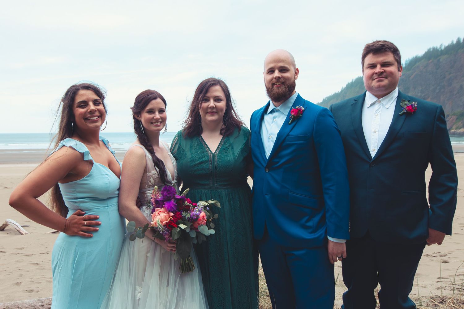Five wedding party members posing together in blue and green attire on a beach.