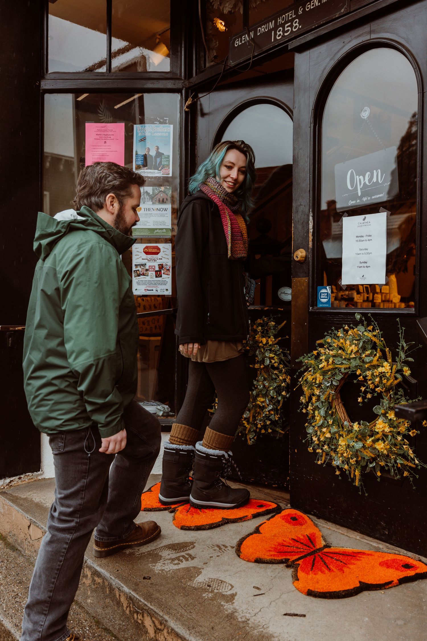 A bearded man holds open a shop door for a smiling blue-haired woman on the steps.