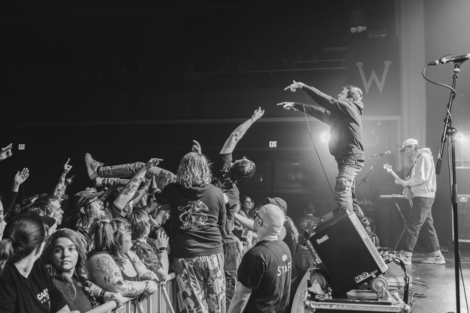 Black and white photo of performer on speaker pointing at cheering crowd during high-energy rock concert.