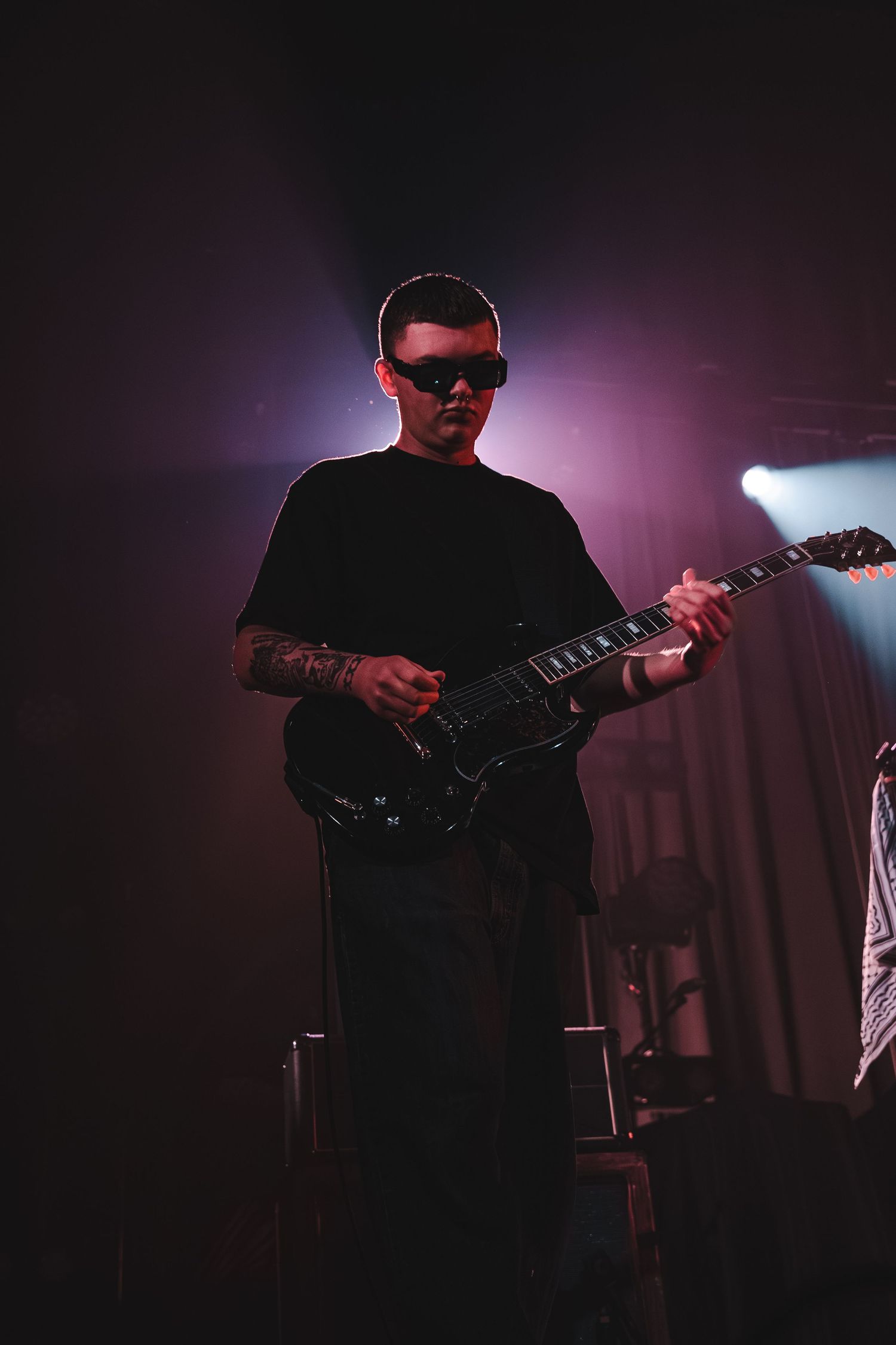 Tattooed guitarist in sunglasses playing black electric guitar on stage under purple and pink stage lighting.