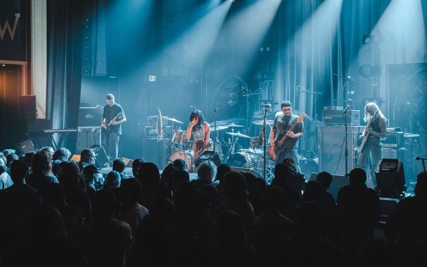 Full band performs on stage under blue spotlights before a large crowd at a live concert.
