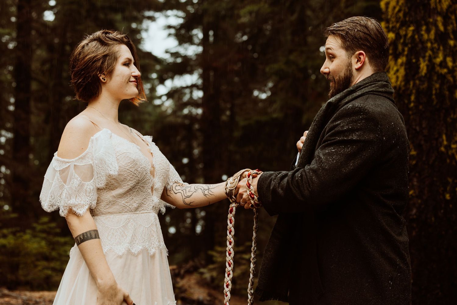 Bride and groom exchange vows with hands bound in colorful handfasting cord during forest wedding.