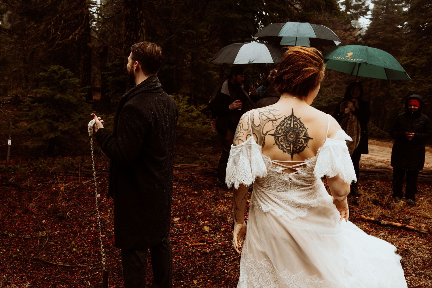 Tattooed bride walks with guests holding umbrellas during a rainy forest wedding ceremony.