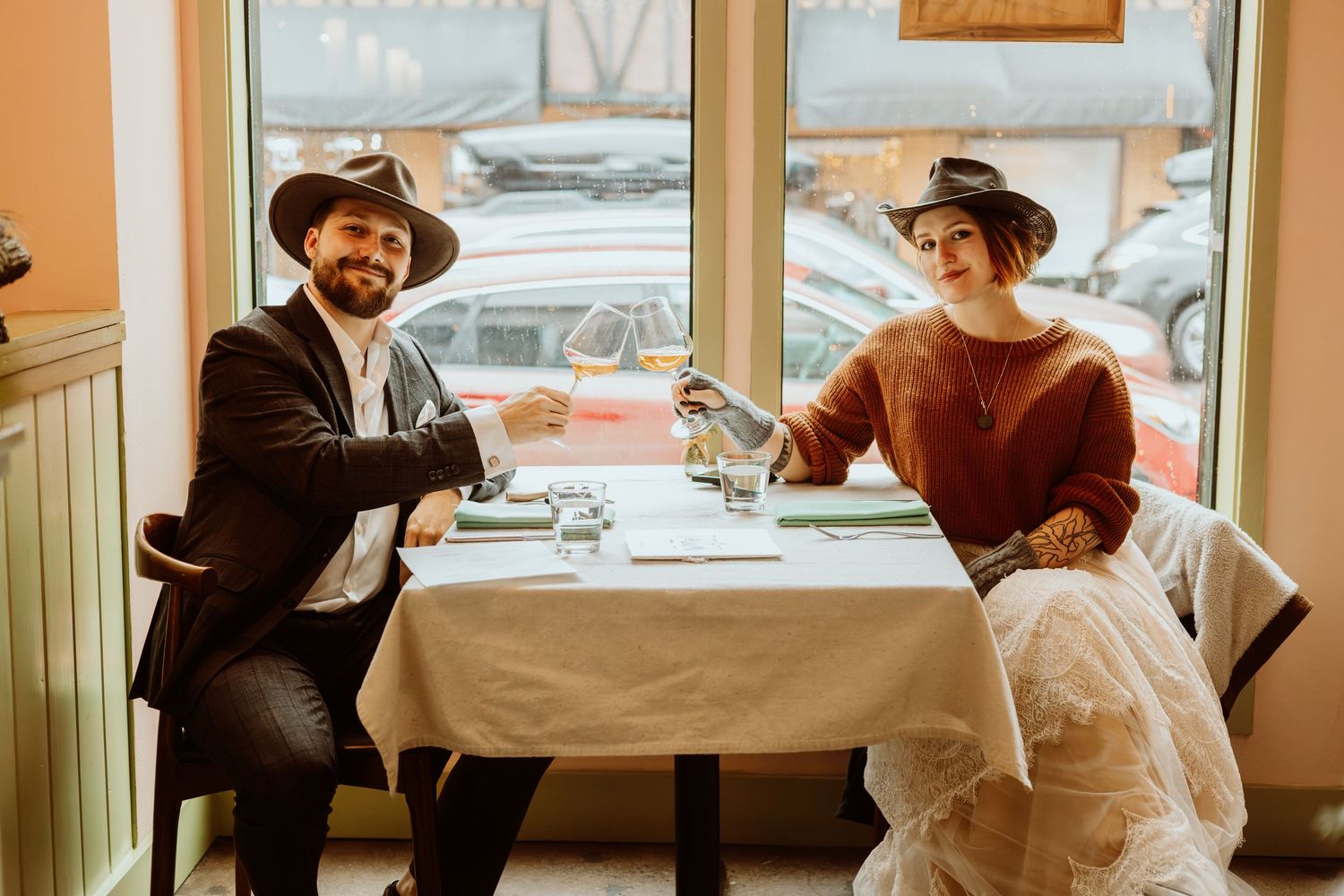 Bride and groom in matching cowboy hats smile while toasting with champagne flutes at an elegant restaurant table.
