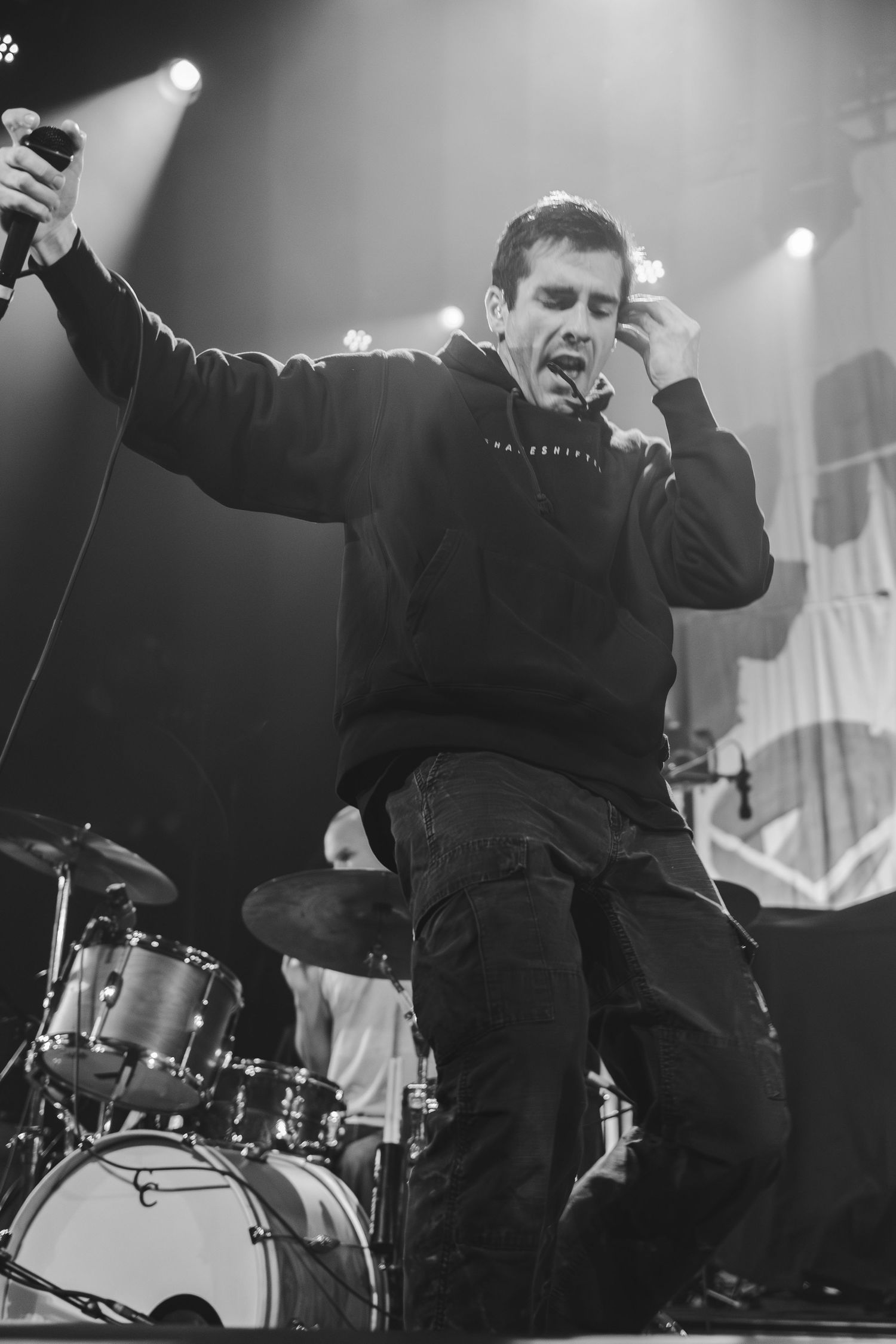 Black and white photo of an energetic male vocalist performing on stage with a drum kit behind him.