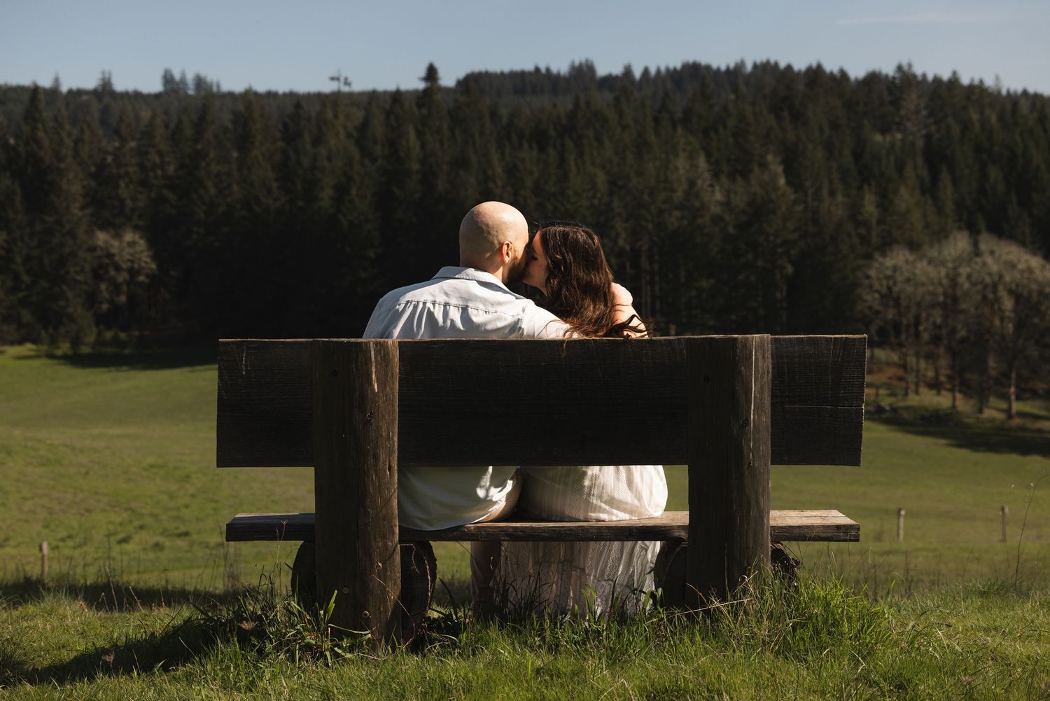 Couple sitting closely together on a wooden bench overlooking a scenic green forest meadow.
