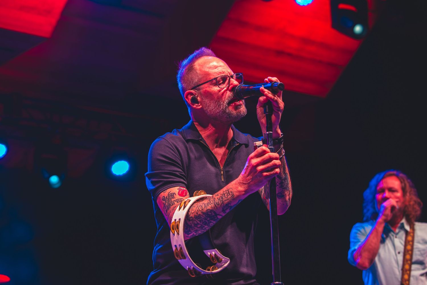 Tattooed male vocalist performs on stage holding a tambourine under blue and red lights.