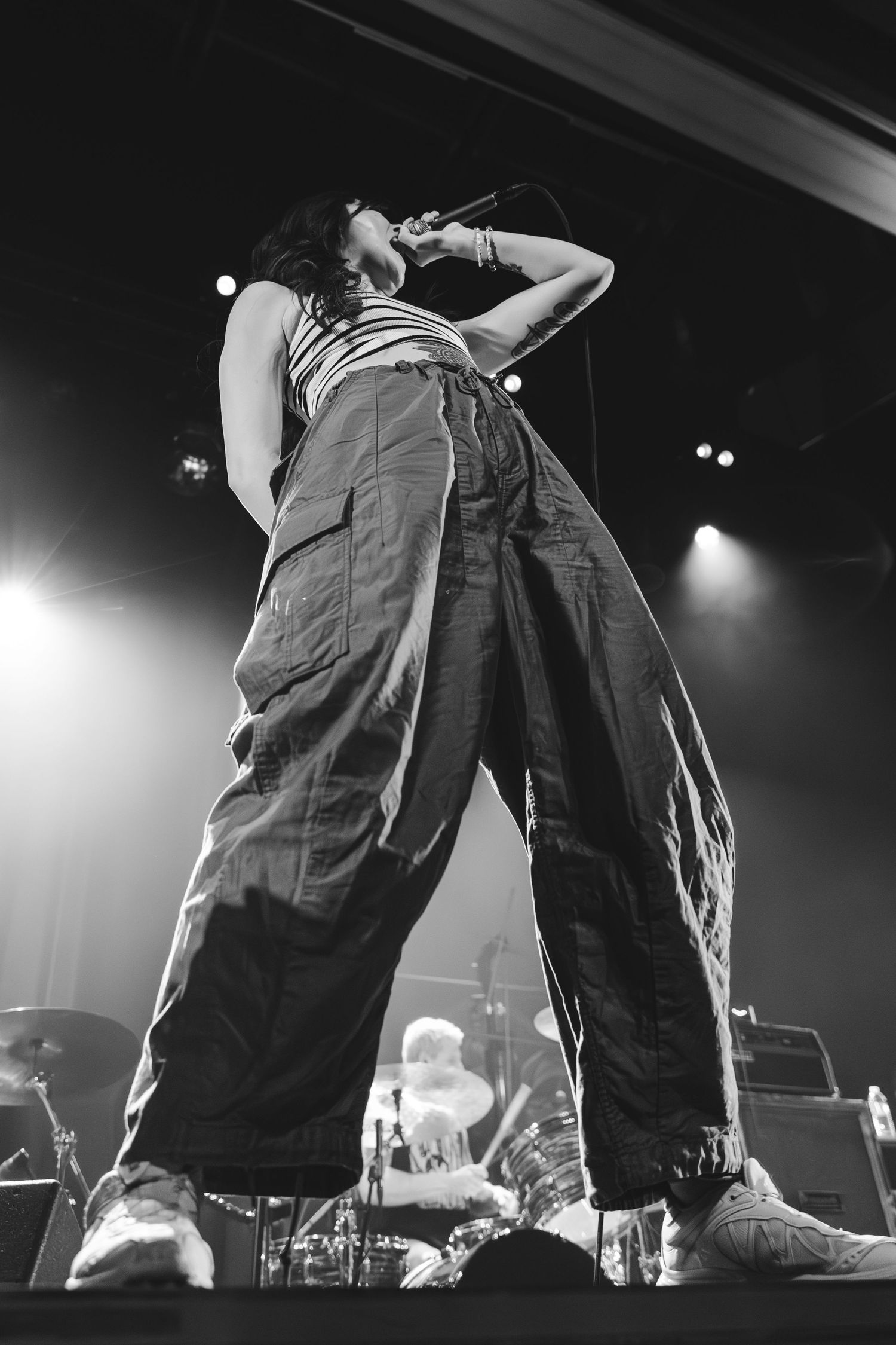 Low angle black and white shot of vocalist performing on stage in baggy pants under bright concert lights.