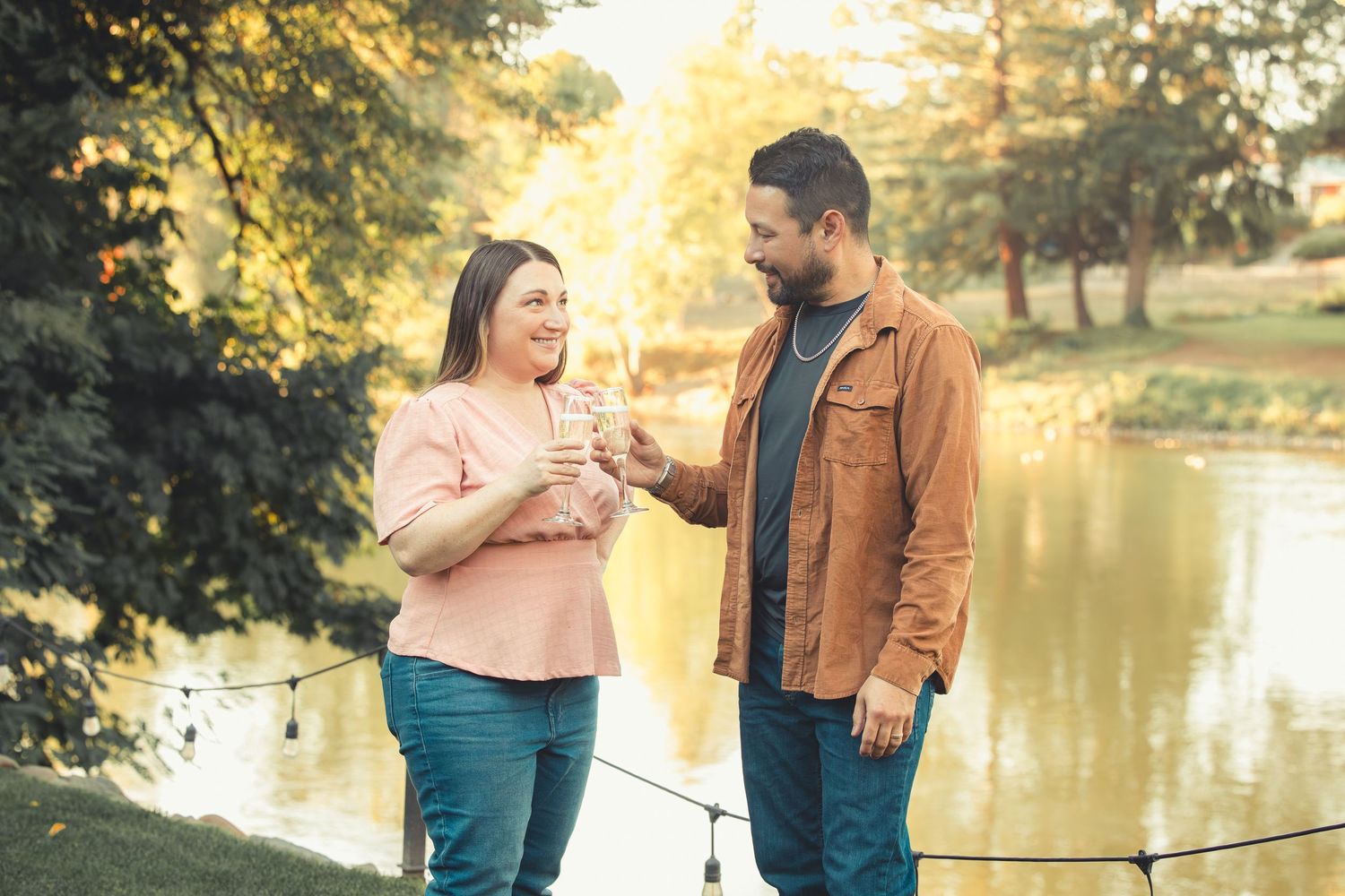 Couple sharing a romantic moment by a sunlit lake, exchanging drinks among string lights and golden trees.