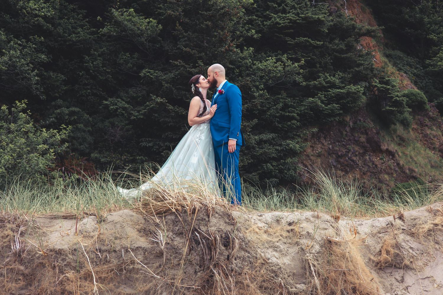 Bride and groom share tender moment on sandy coastal dune surrounded by dense evergreen trees.