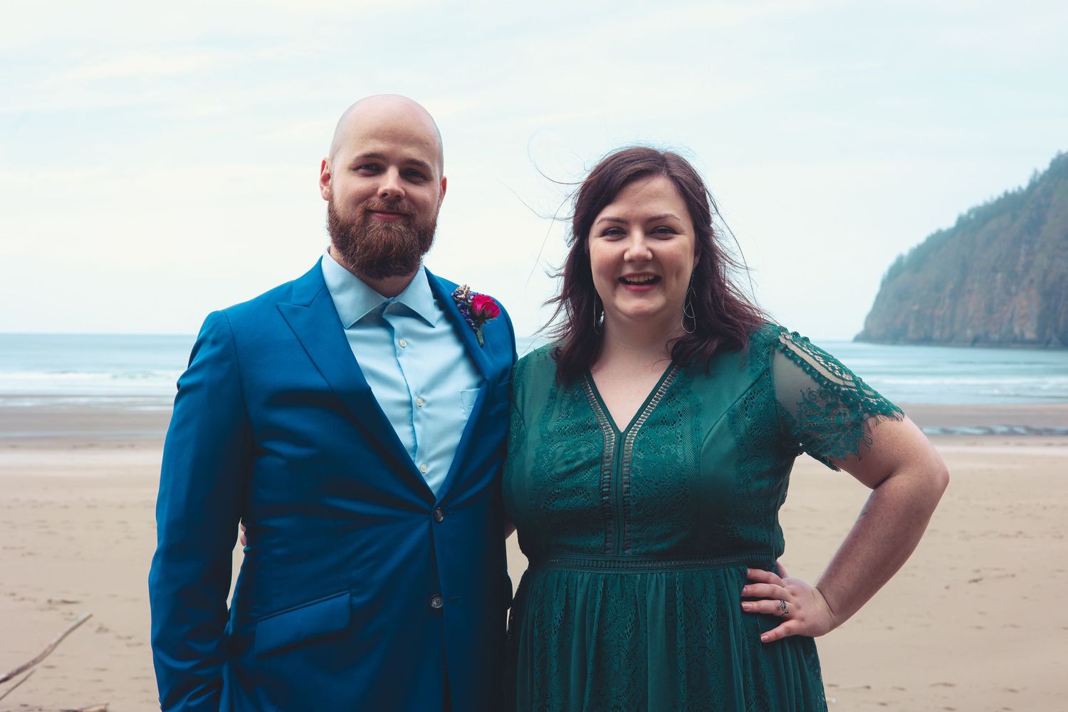 A man in a blue suit and woman in an emerald green dress stand together on a cloudy beach.