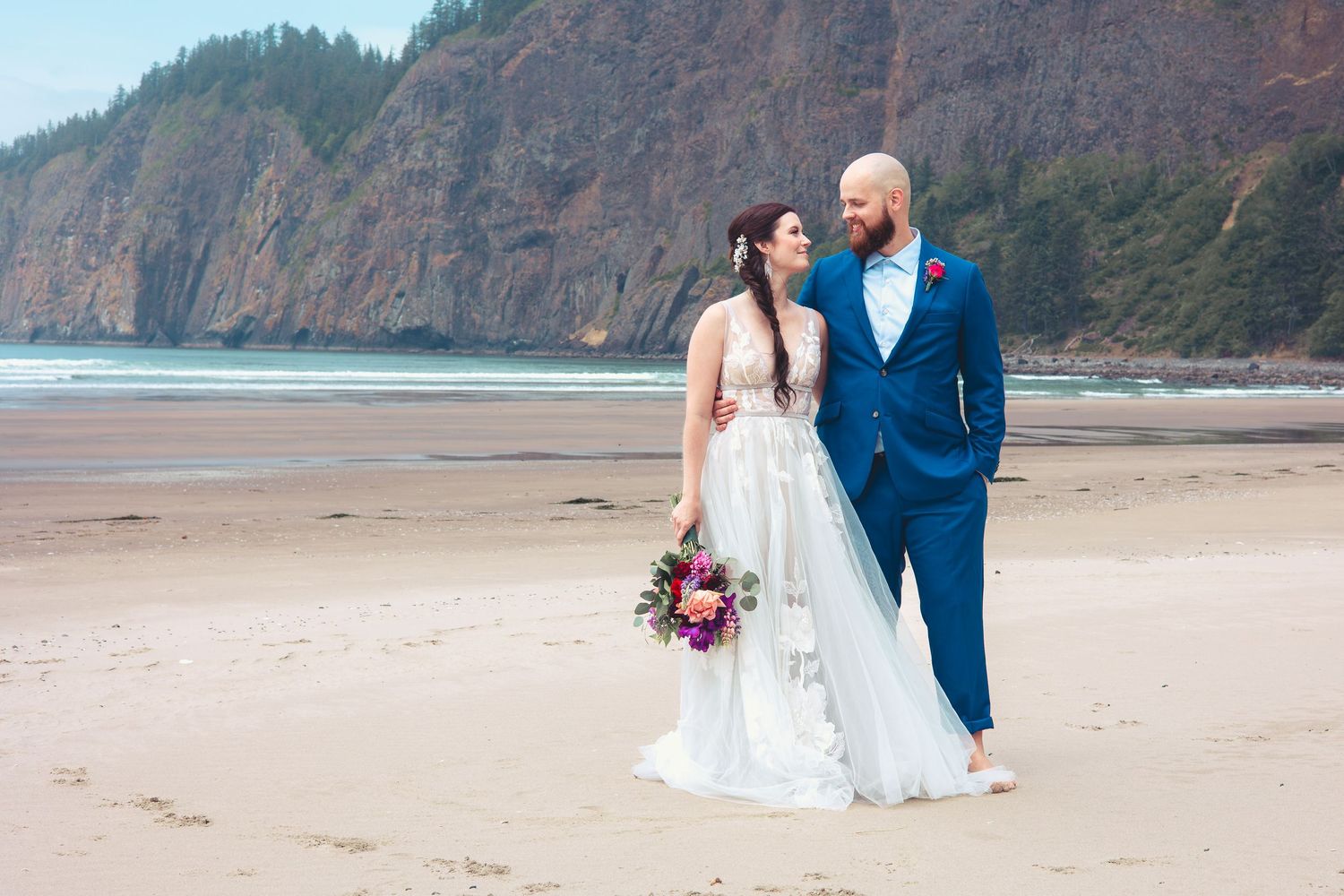 Bride and groom walking on a sandy beach with dramatic cliffs, bride holding a colorful bouquet.