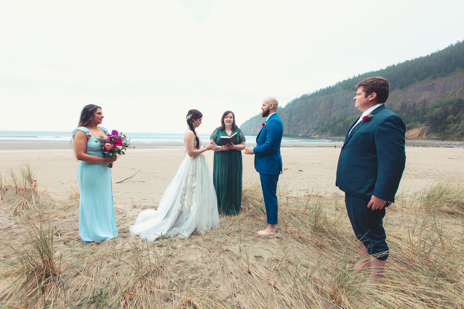 Small beach wedding ceremony with bride, groom, officiant, and two guests on a sandy shoreline.