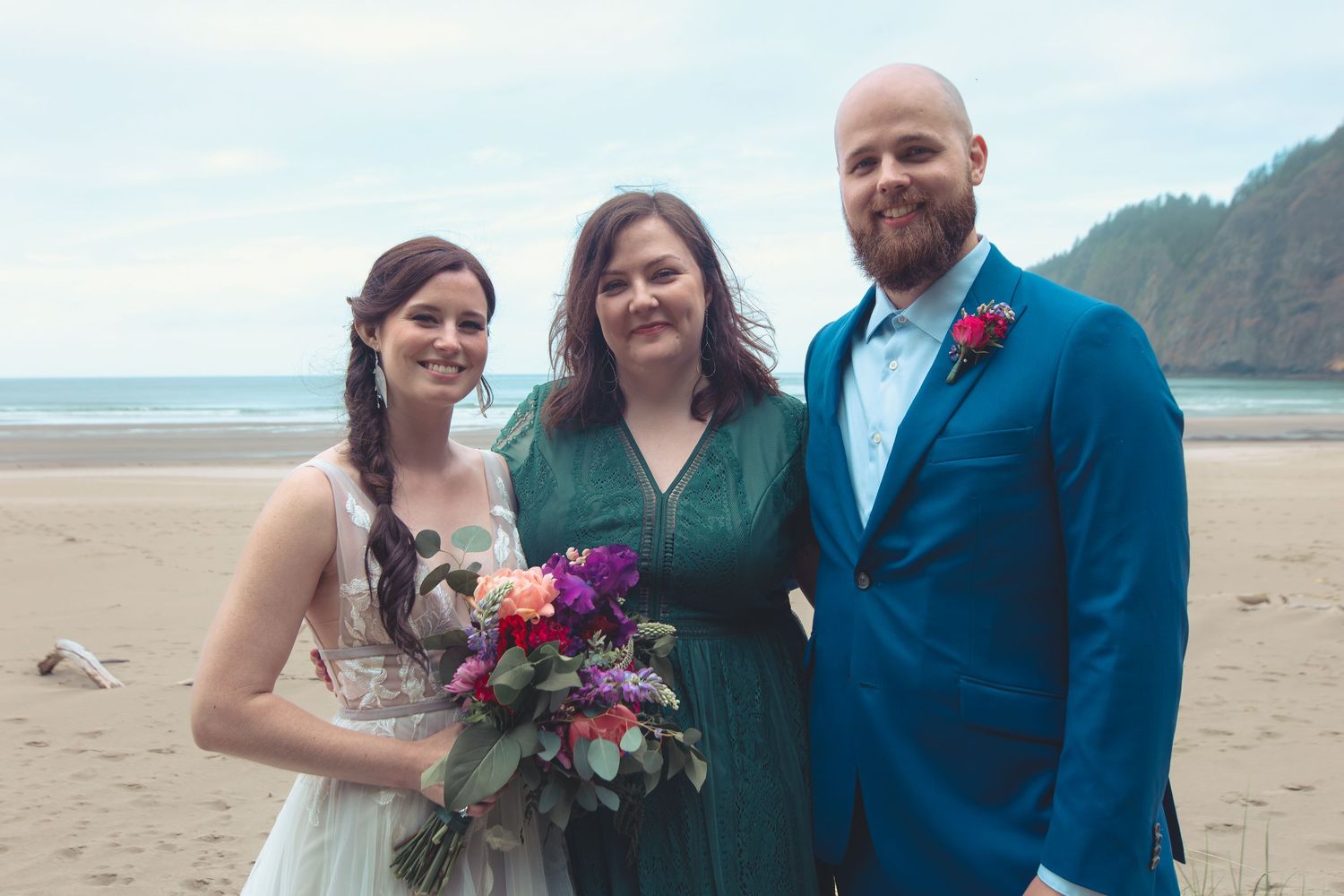 Bride holding colorful bouquet stands with groom and bridesmaid on a beach.