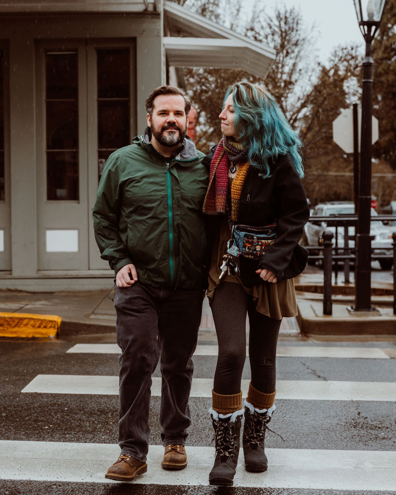 A couple standing on a crosswalk smiling, woman with teal hair wearing colorful scarf.