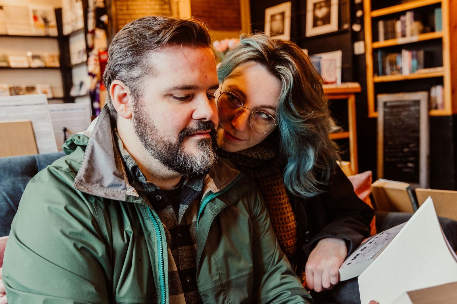 A blue-haired woman leans affectionately on a bearded man reading a book in a bookstore.