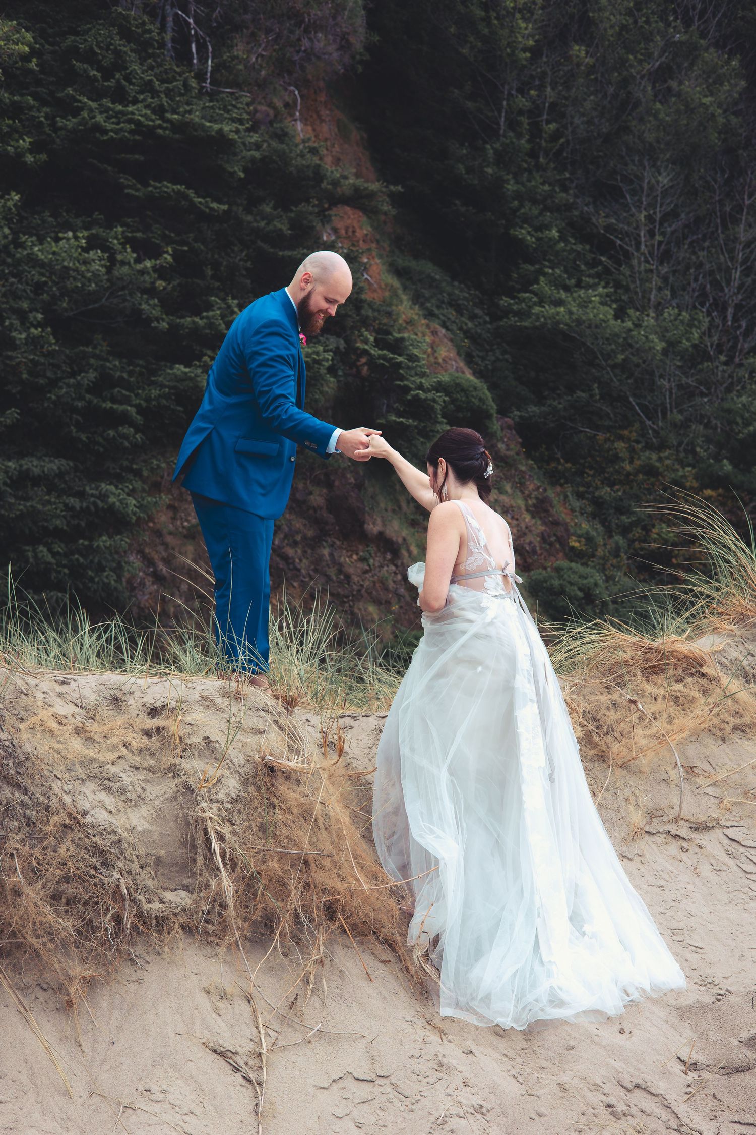 Groom in blue suit helps bride in flowing gown climb sandy dune at beach wedding.