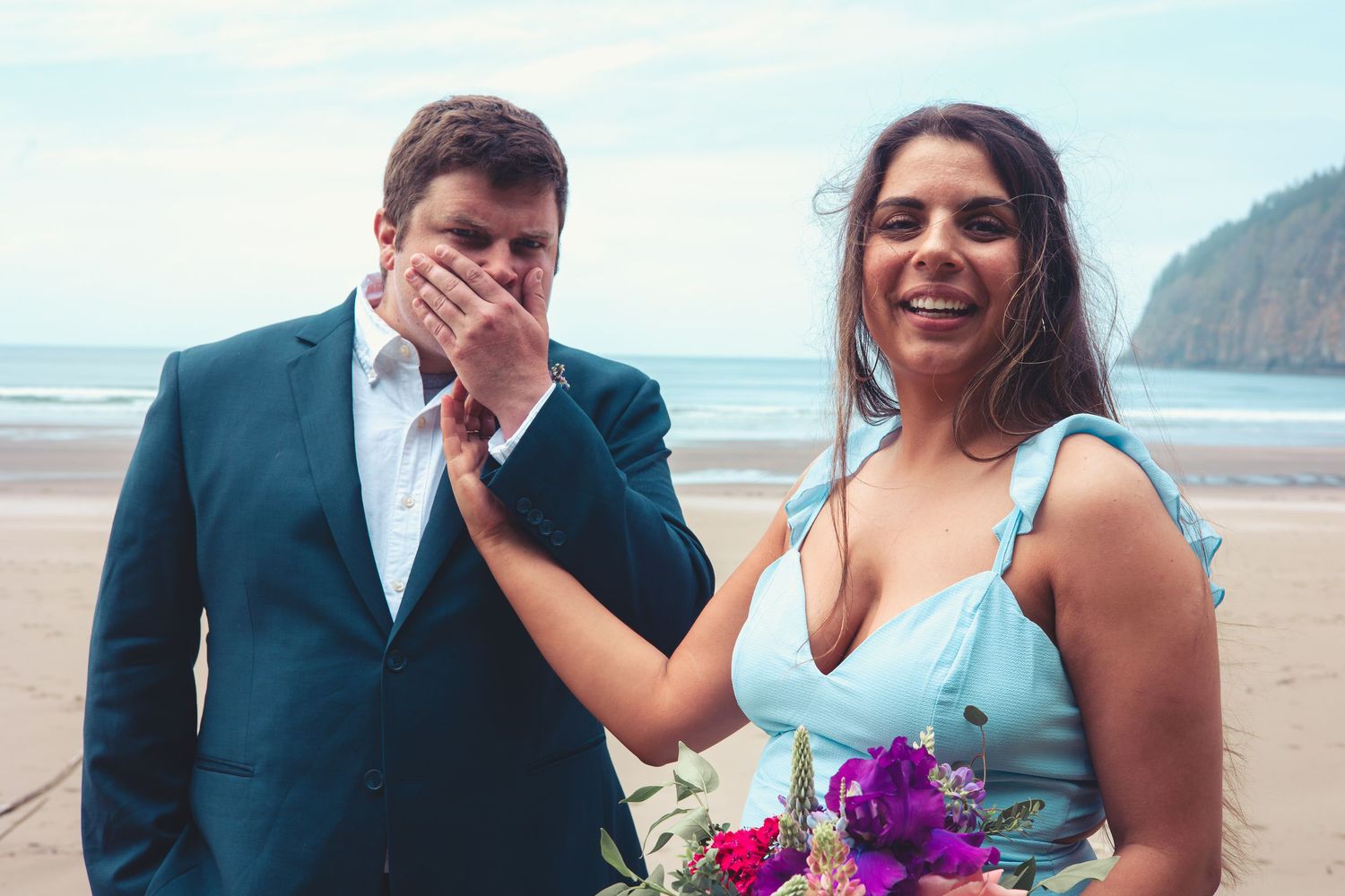 Man in teal suit and smiling woman in blue dress laugh together on a beach.