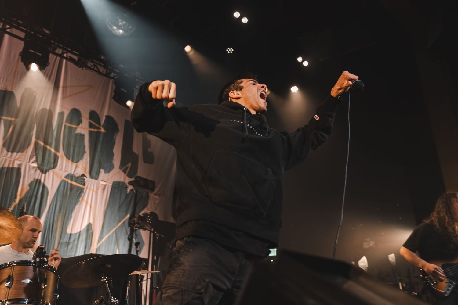 Energetic vocalist in black hoodie raising fists before a large crowd at a live concert.