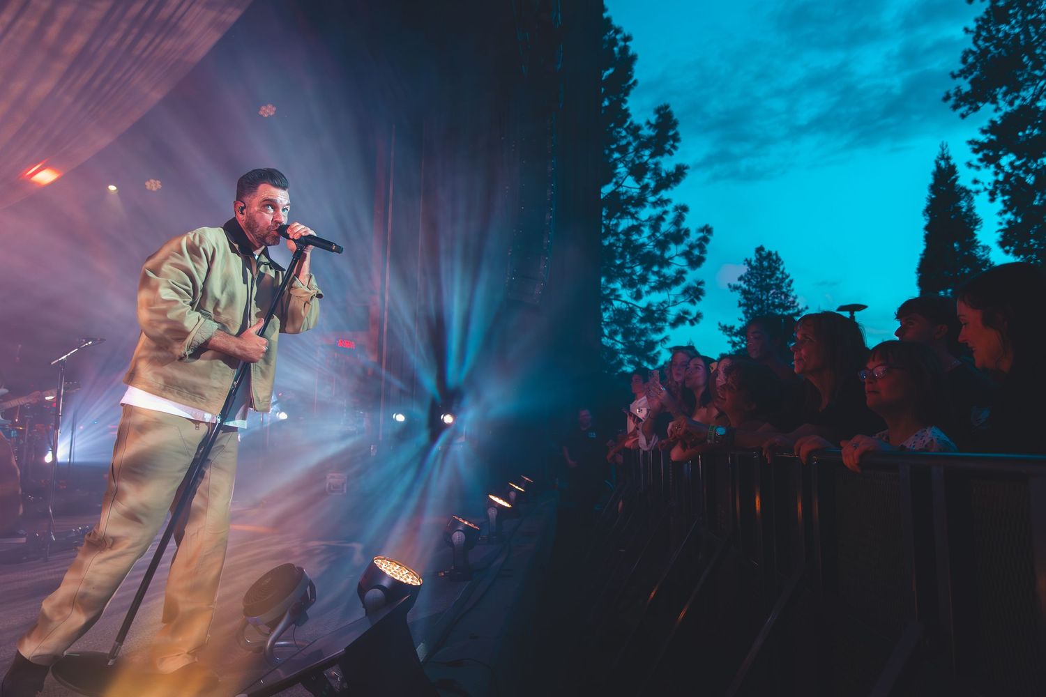 A male singer performs on an outdoor stage at twilight with fans cheering at the front barrier.