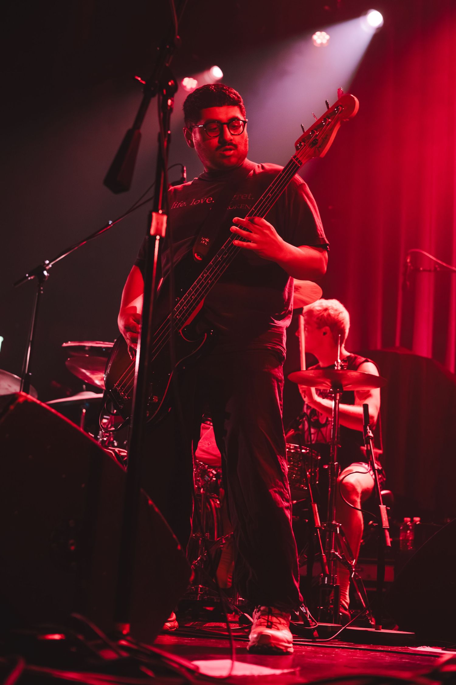 Bassist wearing glasses plays electric bass guitar on stage bathed in red concert lighting.