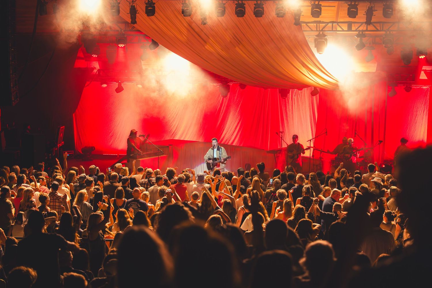 A large crowd enjoys a live concert under red stage lights with draped fabric backdrop.