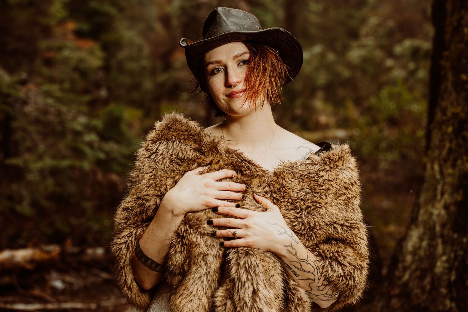 Bride in fur stole and black hat smiling softly, posing among trees in a forest setting.