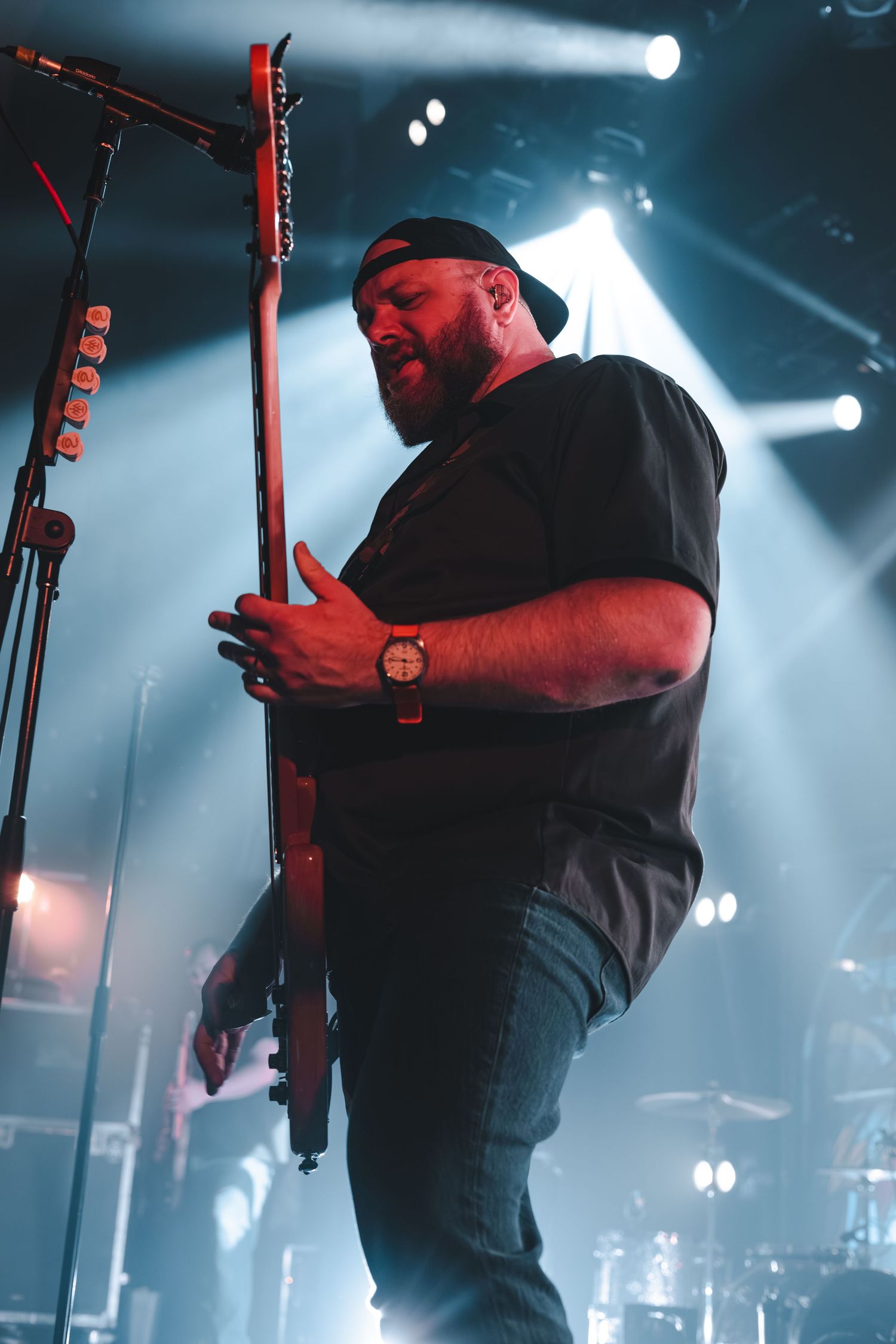 Bearded guitarist in black t-shirt performing passionately on stage under red and blue concert lights.