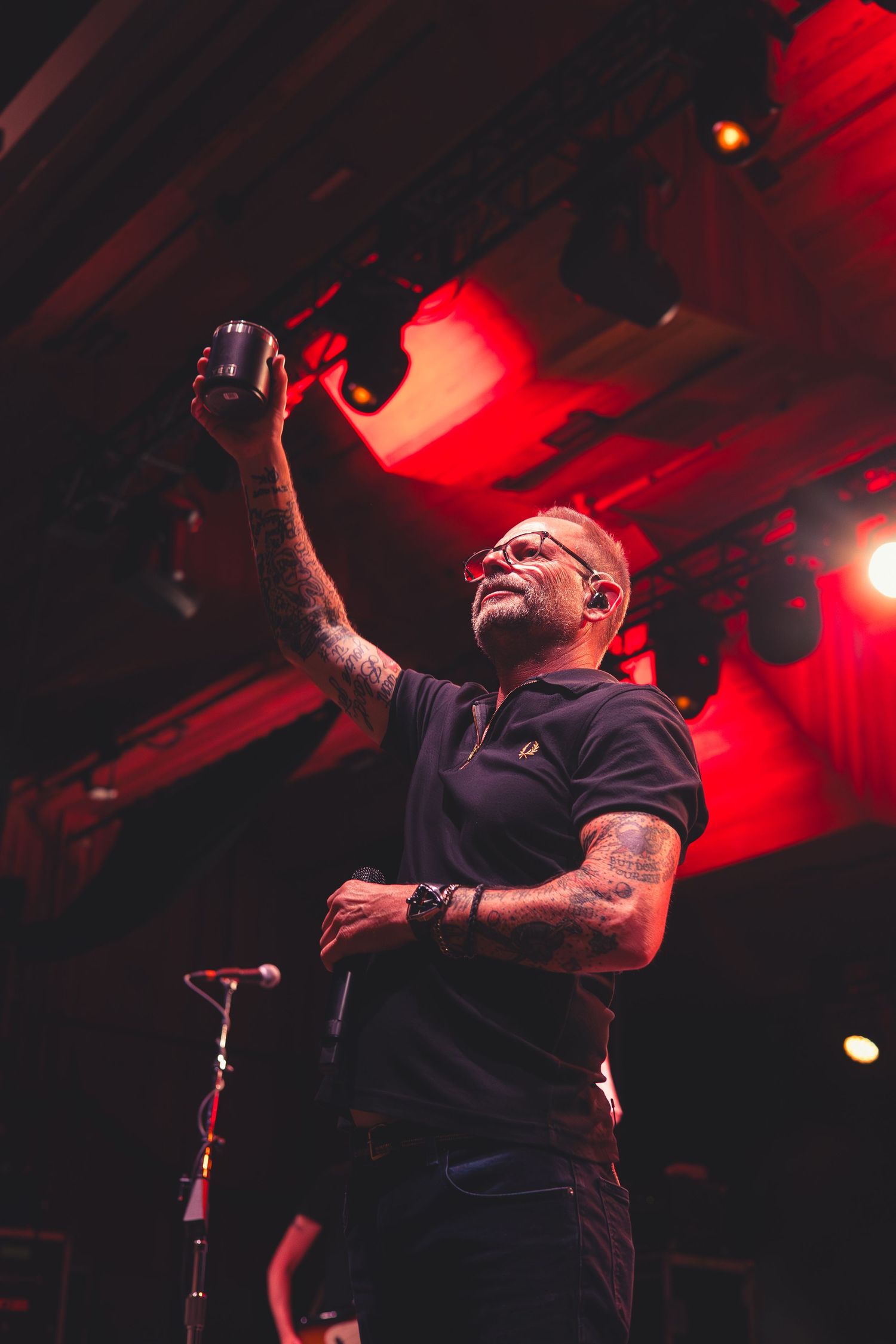 Tattooed male vocalist raising a microphone cup toward the ceiling under dramatic red stage lighting at a concert.