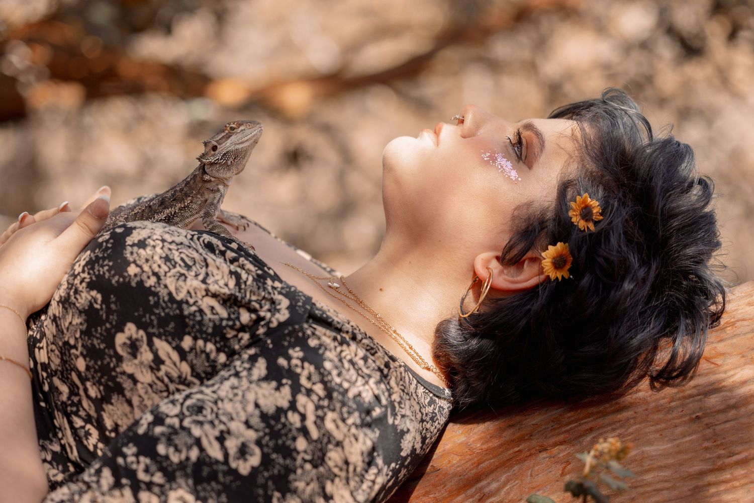 Woman with sunflower hair accessories lies back on a wooden surface with a lizard resting on her chest.